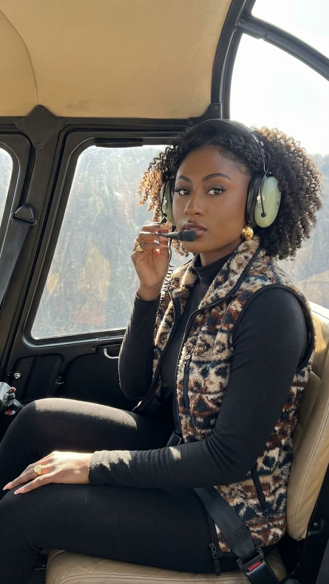A woman wearing headphones and a patterned vest sits in a helicopter seat, holding a pen near her mouth and looking at the camera. Sunlight shines through the window beside her, revealing glimpses of Switzerland’s fairytale villages in winter below.