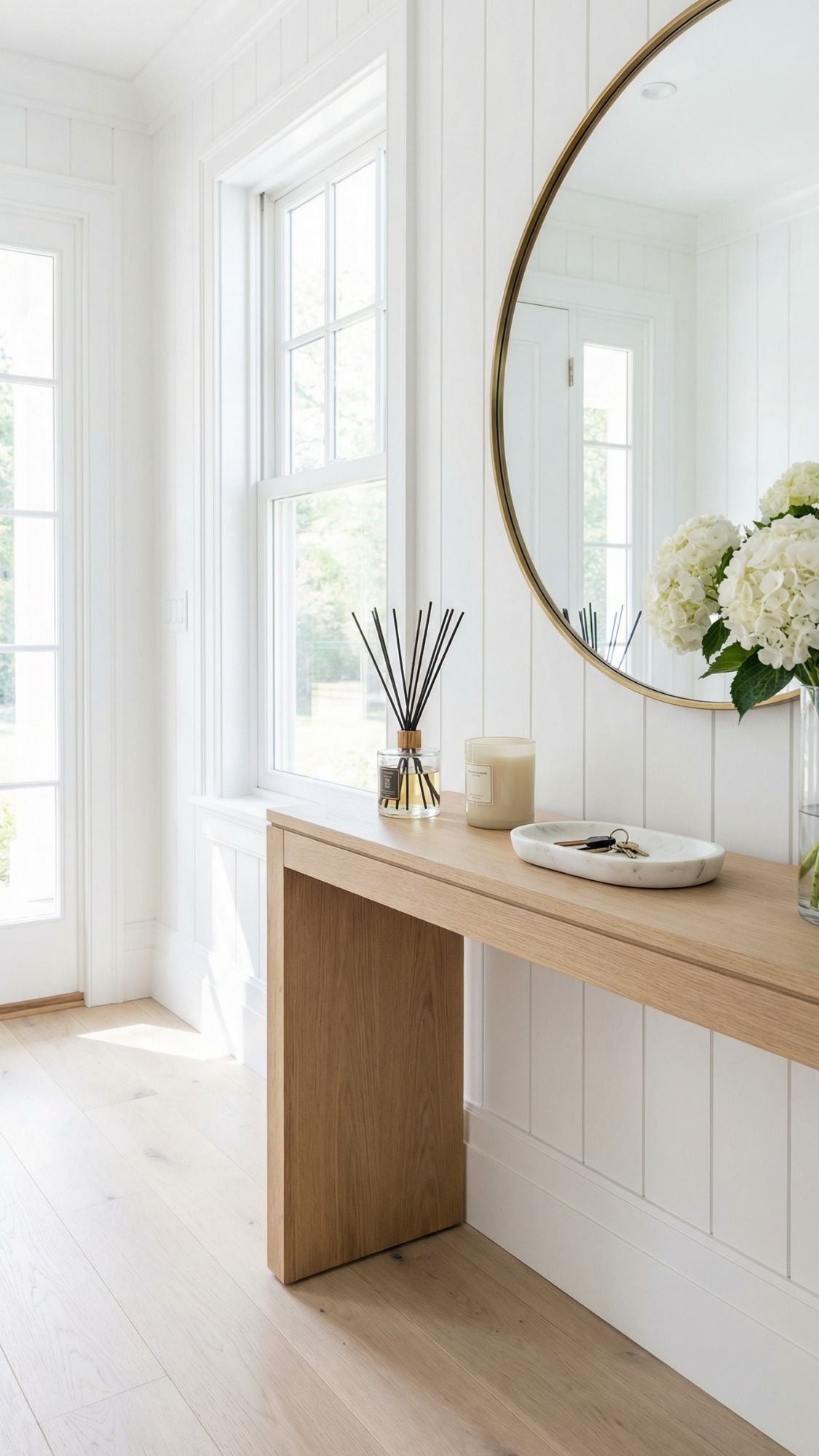 A light-filled entryway with white paneled walls, a large round mirror, and a wooden console table holding a candle, reed diffuser, decorative tray, and a vase of white flowers—perfect spring home decor ideas to refresh your space.