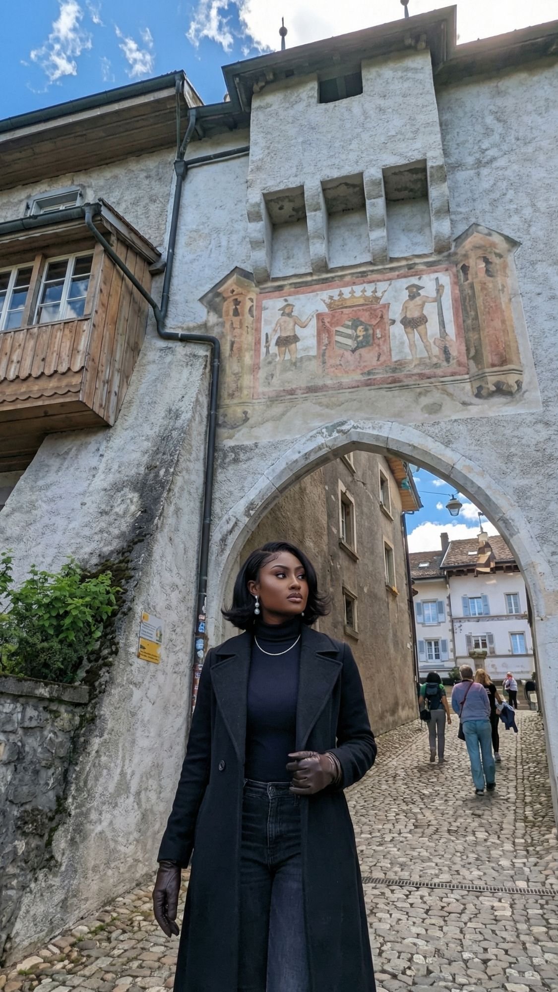 A woman in dark clothing stands under a historic stone archway with a painted mural, surrounded by fairytale villages and old European buildings on cobblestone streets in Switzerland, while several people walk in the wintery background.