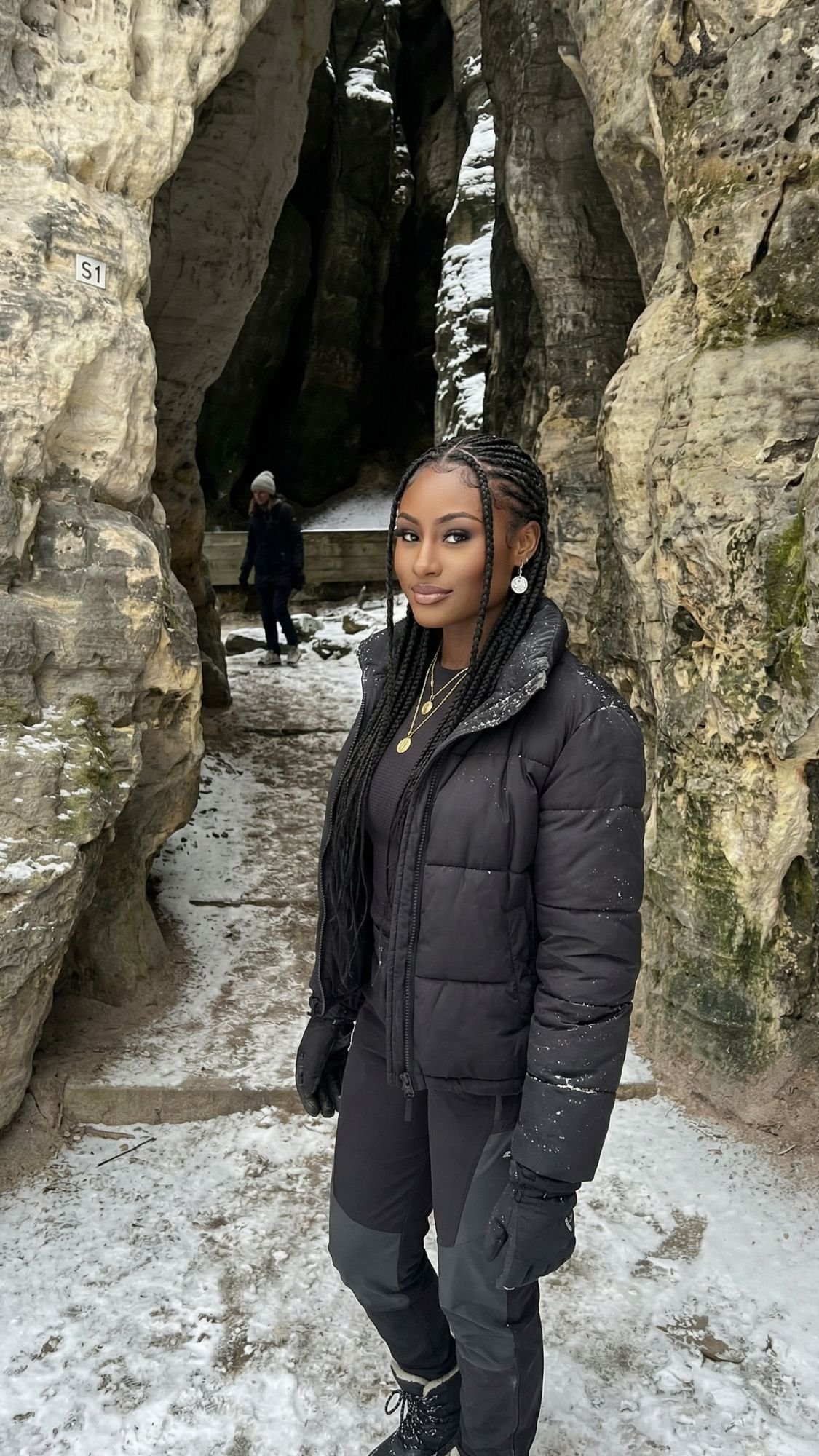 A woman with long braids, wearing a black jacket and gloves, stands on snow between tall rock formations in a winter scene reminiscent of fairytale villages in Switzerland. Another person is in the background near the rocks.