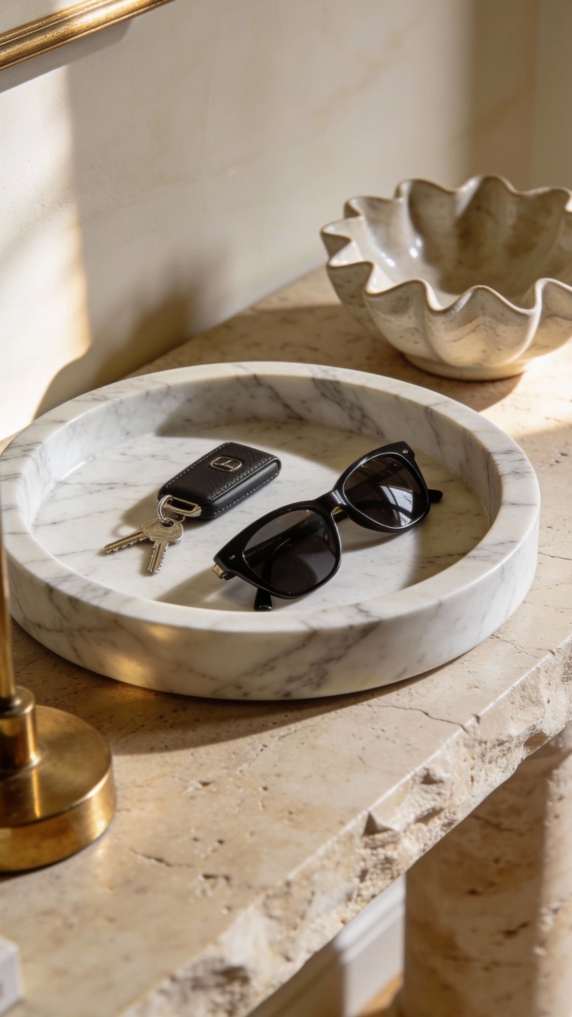 A marble tray on a stone surface holds black sunglasses and keys, with a wavy metallic dish and brass lamp nearby. Sunlight casts soft shadows, offering entryway decor inspiration for warm, welcoming entryways.