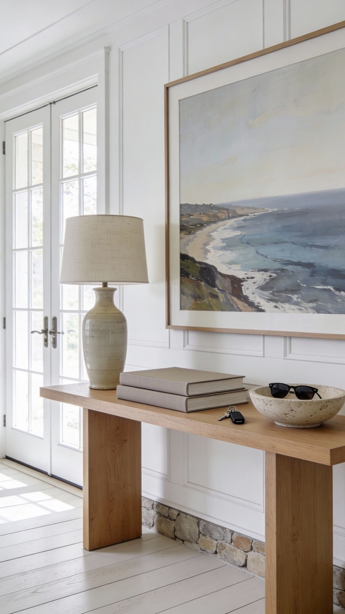 Refresh your space this Spring with a light-filled entryway featuring a wooden console table, ceramic lamp, books, a bowl for sunglasses and keys, and a large framed coastal landscape painting on a white paneled wall.