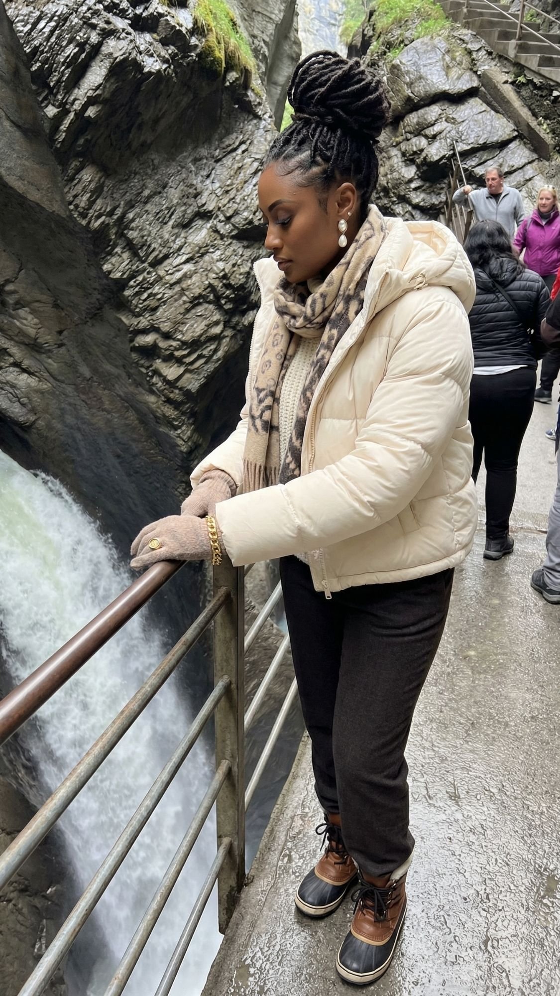A woman in a white puffer jacket, scarf, and boots stands by a metal railing overlooking a rocky waterfall in Switzerland, with other people strolling behind her through this scenic, winter landscape reminiscent of fairytale villages.