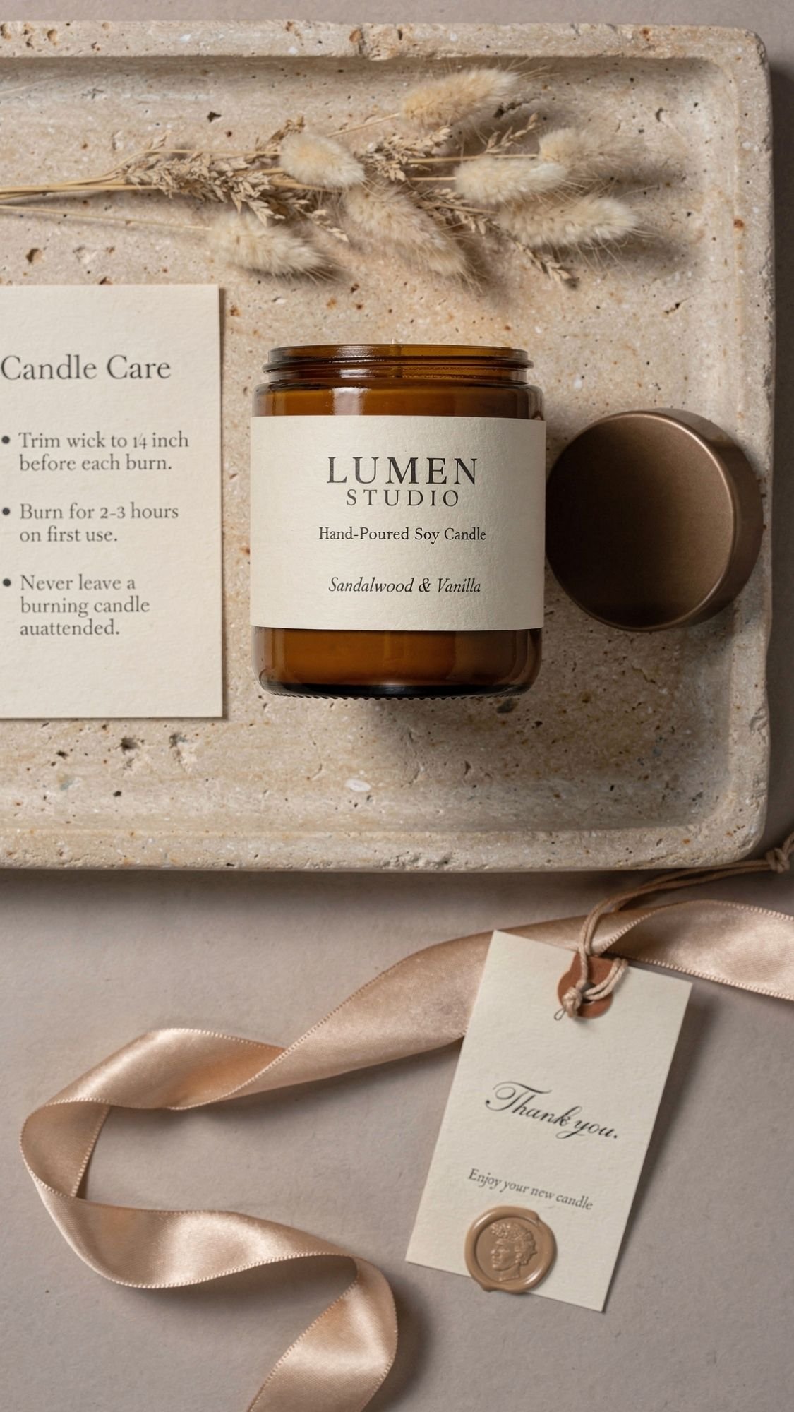 A brown glass jar candle labeled Lumen Studio Sandalwood & Vanilla sits on a speckled tray with a dried floral arrangement, an open lid, neutral aesthetic packaging, a candle care card, and a thank you tag attached to a satin ribbon.