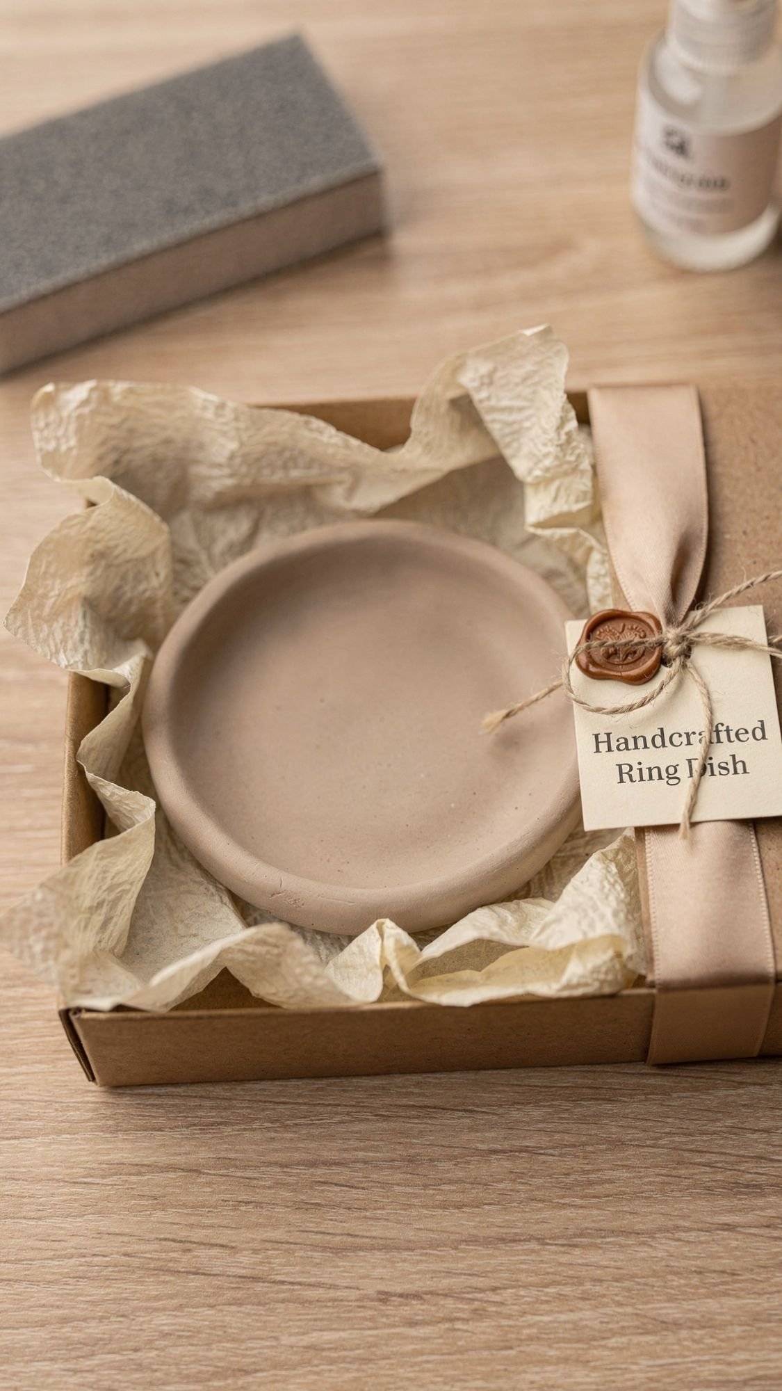 A beige handcrafted ring dish rests on crinkled tissue paper inside an open gift box, showcasing neutral aesthetic packaging. A tag reading Handcrafted Ring Dish is tied with twine and sealed with wax—perfect for DIY presents or thoughtful gifts for mom.