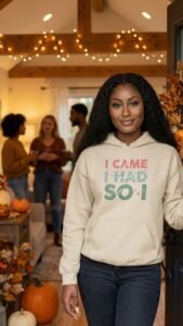 A woman with long curly hair stands smiling at a doorway, wearing a hoodie that says I CAME I HAD SOI. Behind her, four people are gathered in a warmly lit, autumn-themed living room—perfect for an introvert’s cozy Fall Reset and recharge.