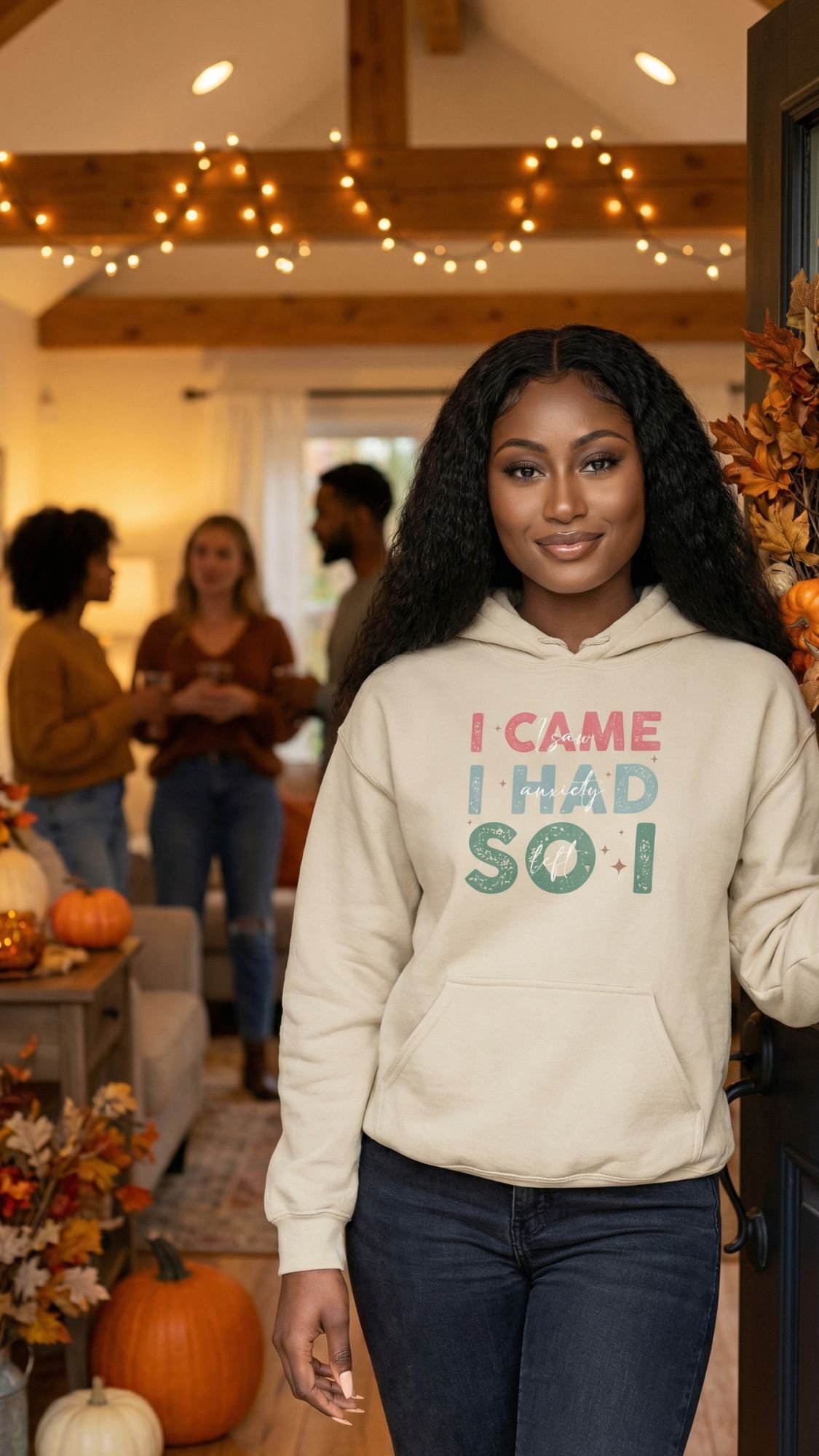 A woman with long curly hair stands smiling at a doorway, wearing a hoodie that says I CAME I HAD SOI. Behind her, four people are gathered in a warmly lit, autumn-themed living room—perfect for an introvert’s cozy Fall Reset and recharge.