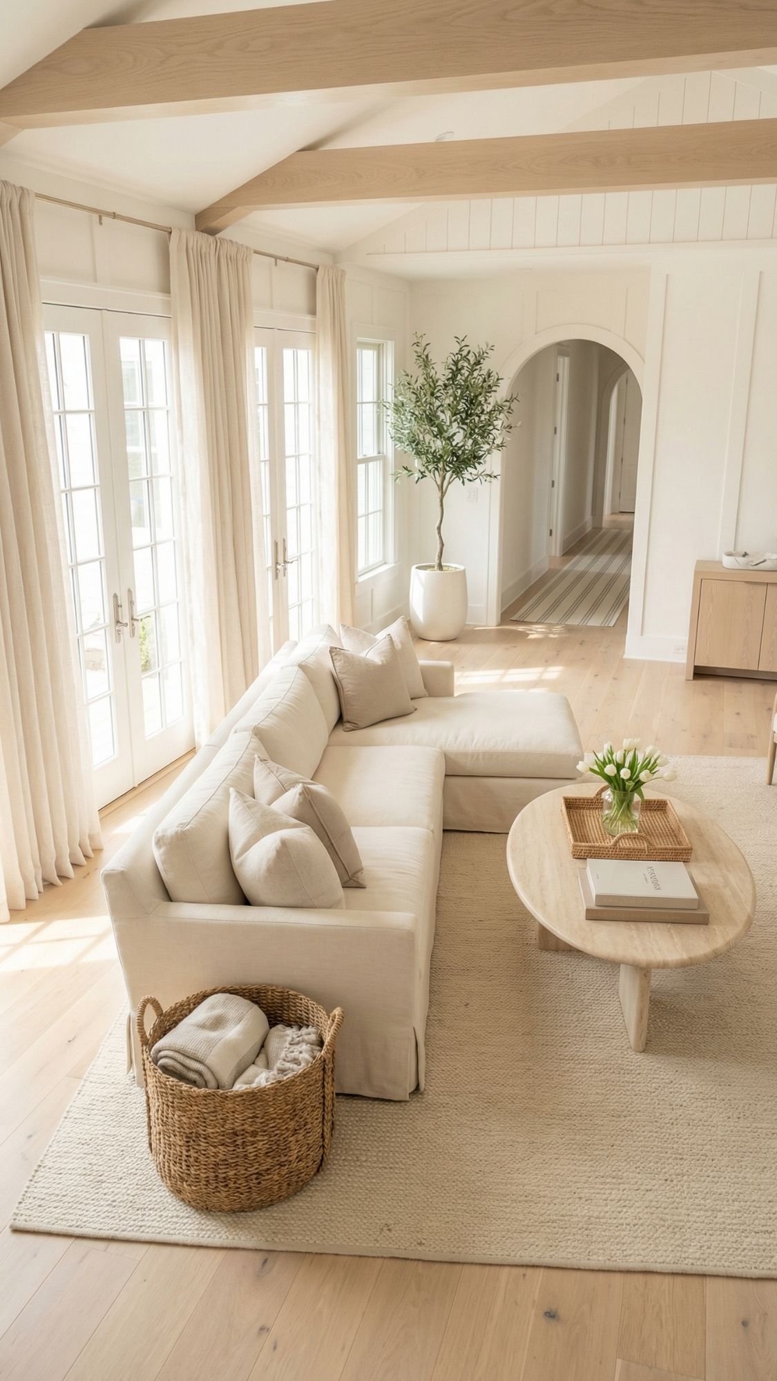 A bright living room with a cream sectional sofa, round wooden coffee table, light rug, and a basket of blankets. Large windows and neutral decor create an airy vibe—perfect home decor ideas to refresh your space this spring.