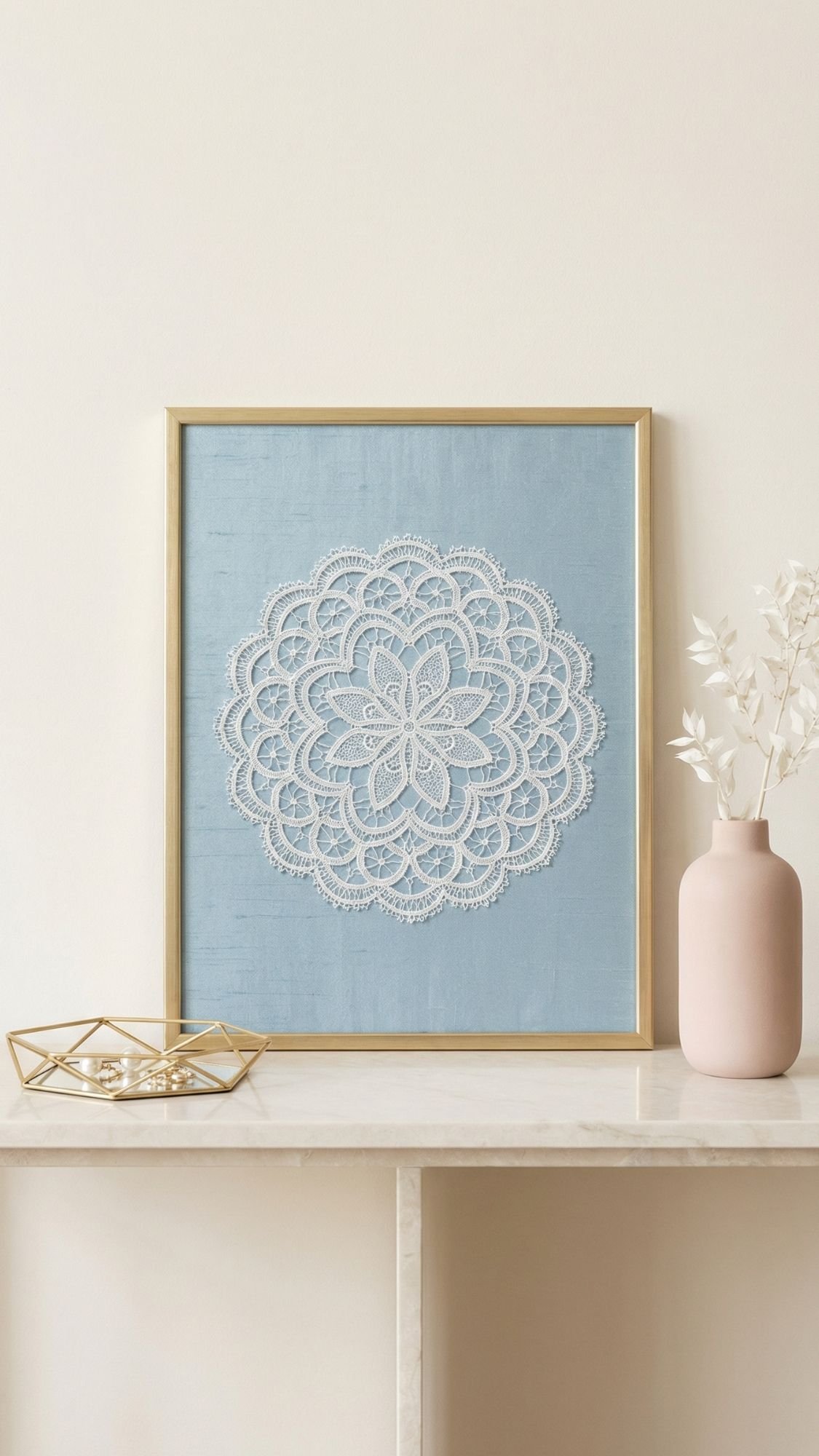 A framed artwork featuring a white lace mandala on a blue background sits on a marble shelf—an elegant idea for DIY Mother’s Day gifts. Nearby are a gold geometric tray and a pink vase with white dried flowers, set against a light-colored wall.
