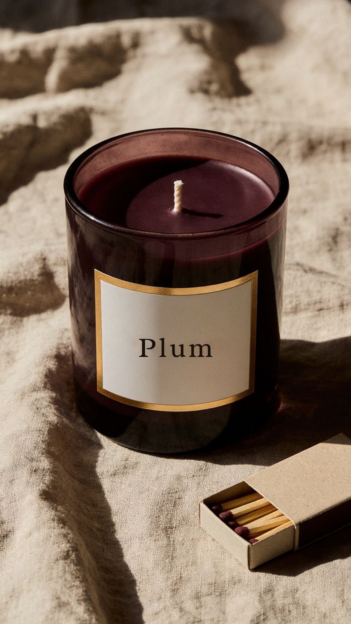 A dark purple candle labeled Plum sits on a beige fabric surface next to an open matchbox filled with red-tipped matches, making it a charming option for handmade gift ideas.
