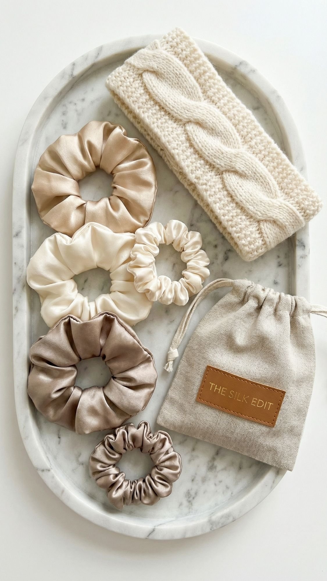A marble tray holds five silk scrunchies in neutral tones, a knitted cream headband, and a beige drawstring pouch labeled THE SILK EDIT—perfect DIY gifts for new moms.