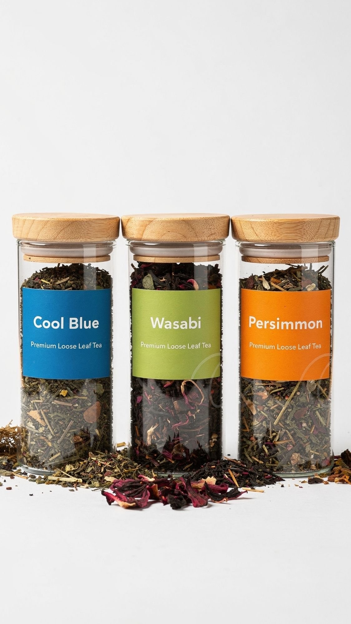 Three glass jars with wooden lids contain loose leaf tea—perfect as best friend gifts. Each jar has a colorful label: blue for Cool Blue, green for Wasabi, and orange for Persimmon. Tea leaves and petals are scattered in front.
