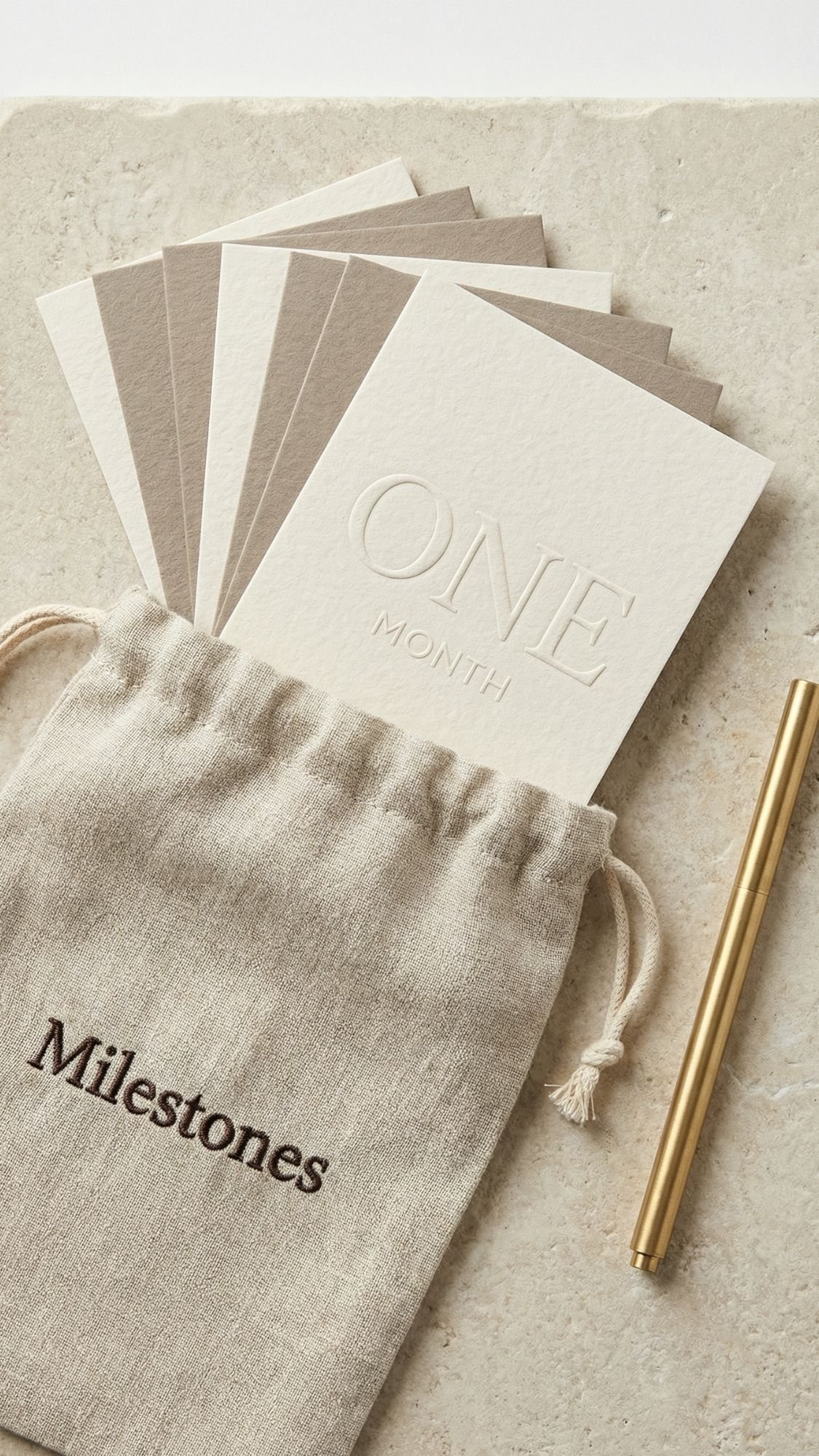 A set of milestone cards, with the top card reading ONE MONTH, sits partially inside a linen pouch labeled Milestones. A gold pen lies beside the pouch on a textured stone surface—gentle gifts and useful gifts for new moms.