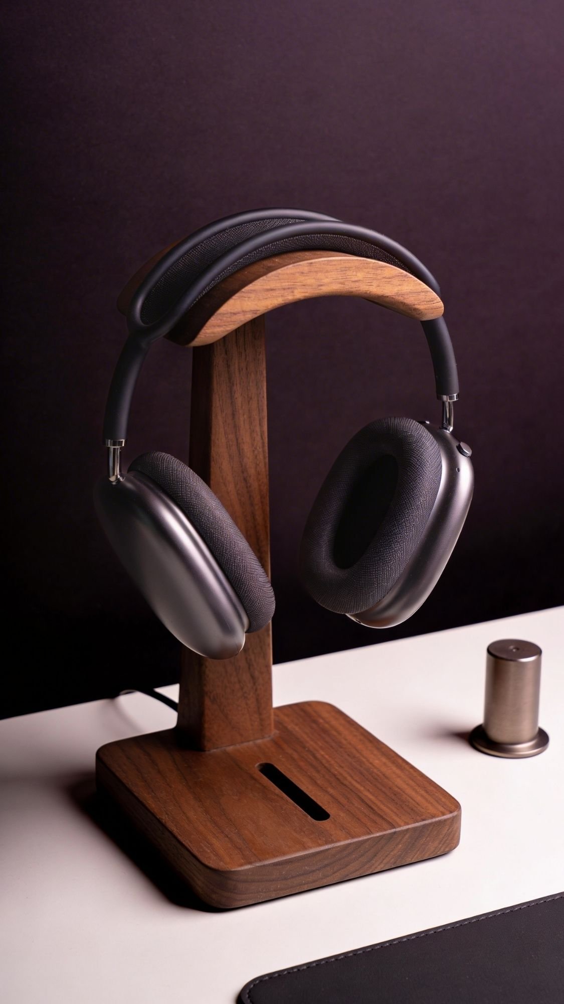 A pair of black over-ear headphones rests on a wooden headphone stand atop a white desk, with a small metallic cylindrical object nearby—an ideal choice for Useful Gifts in a dark, uncluttered setting.