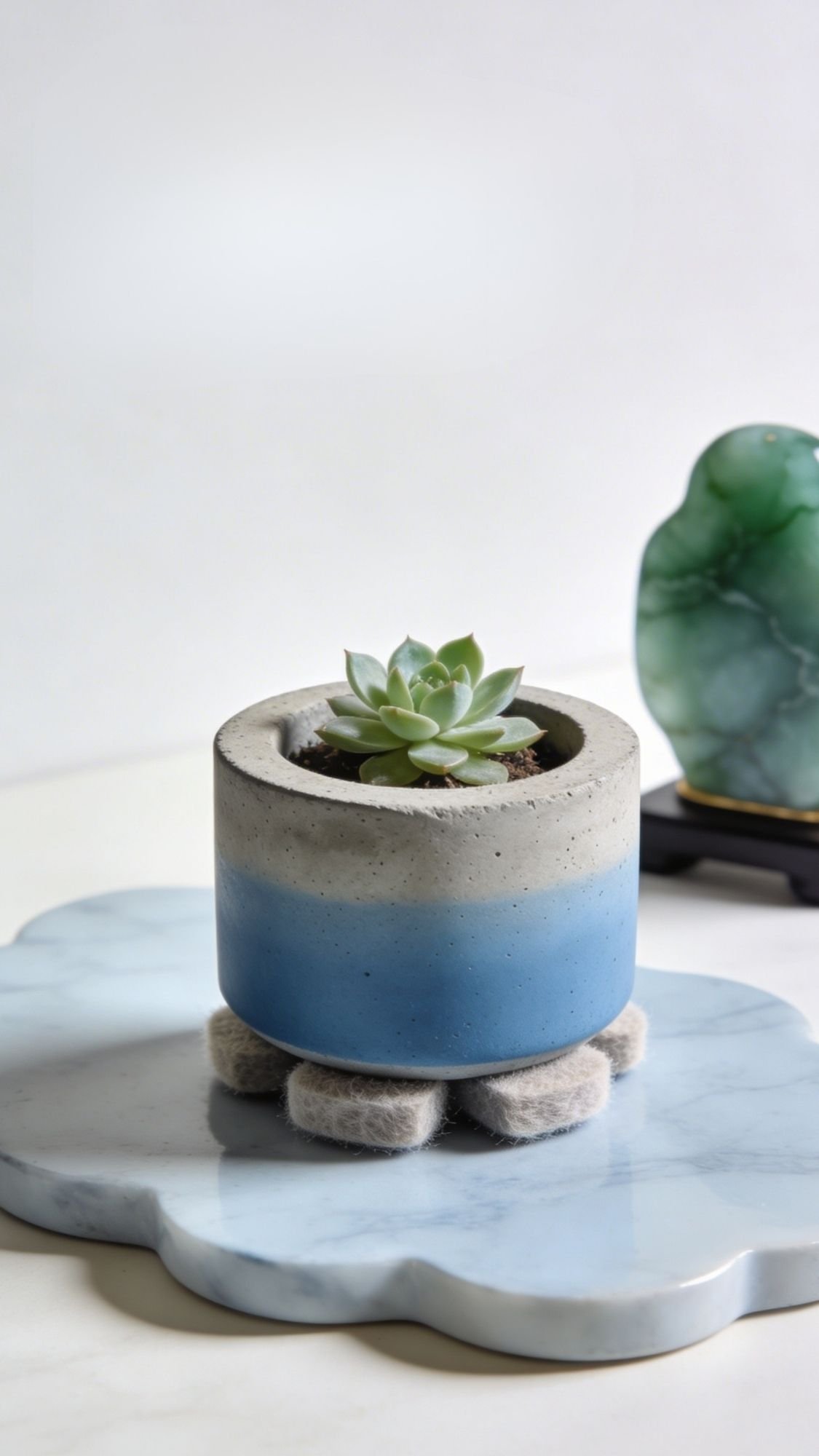 A small succulent in a round, gray and blue concrete planter sits on felt pads atop a cloud-shaped marble tray—an ideal choice for easy handmade gifts or unique birthday gifts for best friend. A green decorative stone object is blurred in the background.