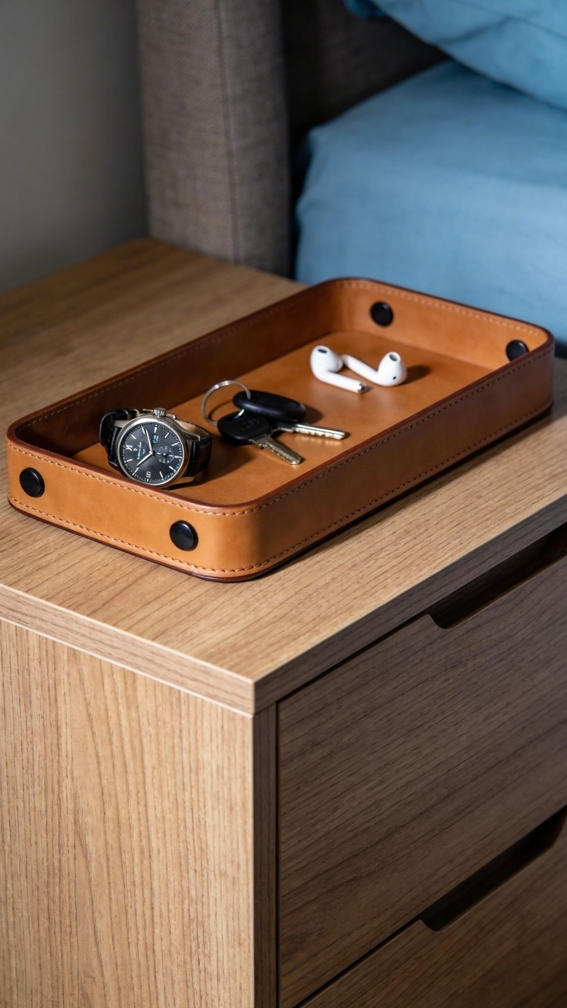 A brown leather tray on a wooden nightstand holds a wristwatch, keys, and wireless earbuds—an ideal choice for Useful Gifts. The nightstand sits by a bed with blue bedding, adding charm and practicality to any space.
