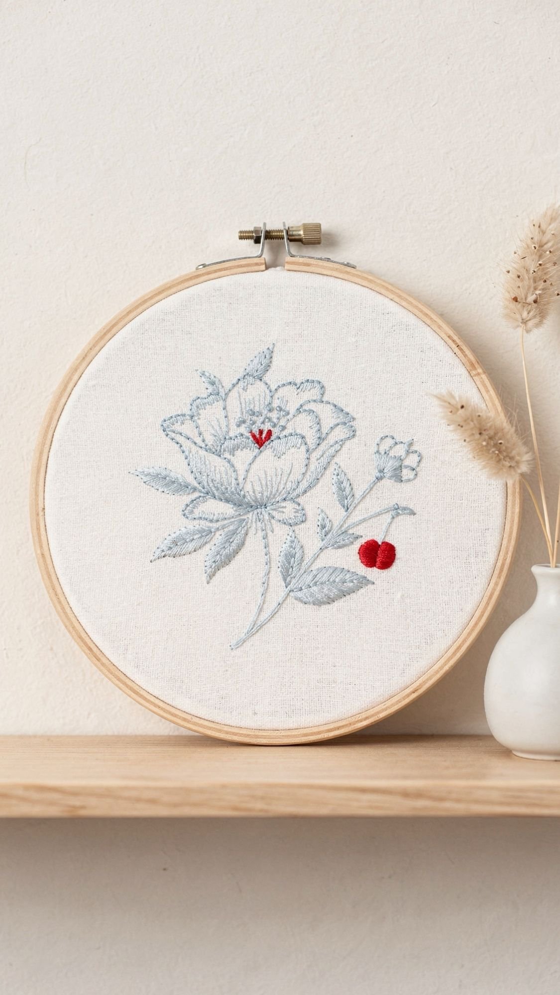 A wooden embroidery hoop displays delicate blue and red floral embroidery on white fabric—a charming idea for DIY gifts. The hoop rests on a light wooden shelf next to a small white vase holding dried grasses.