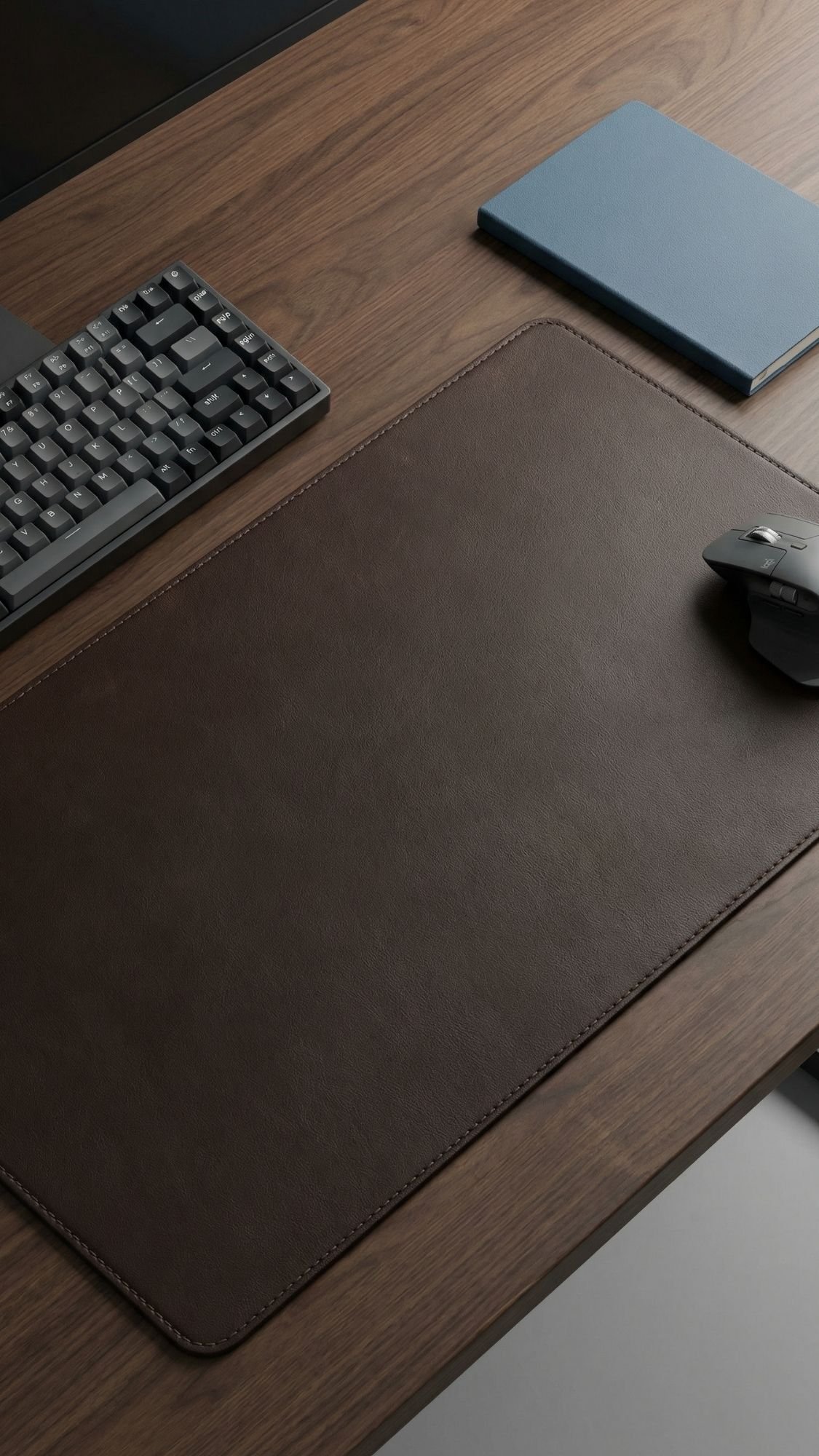 A tidy wooden desk with a black keyboard, a blue closed notebook, a wireless mouse, and a large brown leather desk pad—perfect for those searching for useful handmade gifts or thoughtful gifts for boyfriend.