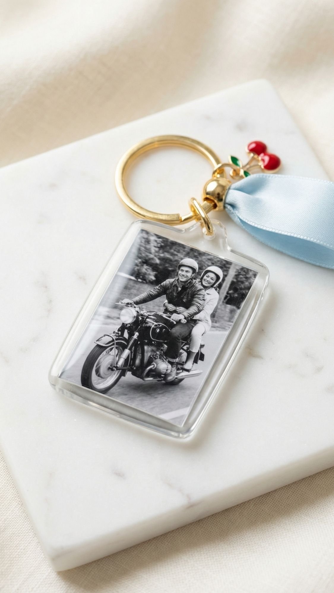 A gold keyring with a blue ribbon and cherry charms holds a clear photo keychain displaying a black-and-white picture of two people riding a motorcycle—perfect for Mother’s Day or as inspiration for heartfelt DIY presents.