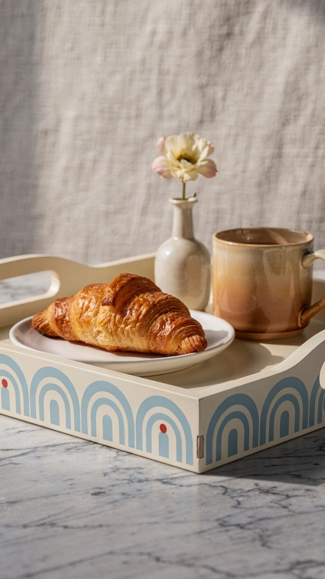 A breakfast tray with a croissant on a white plate, a beige mug, and a small vase holding a single light flower sits on marble—a thoughtful DIY present perfect for Mother’s Day, set against a soft, neutral background.
