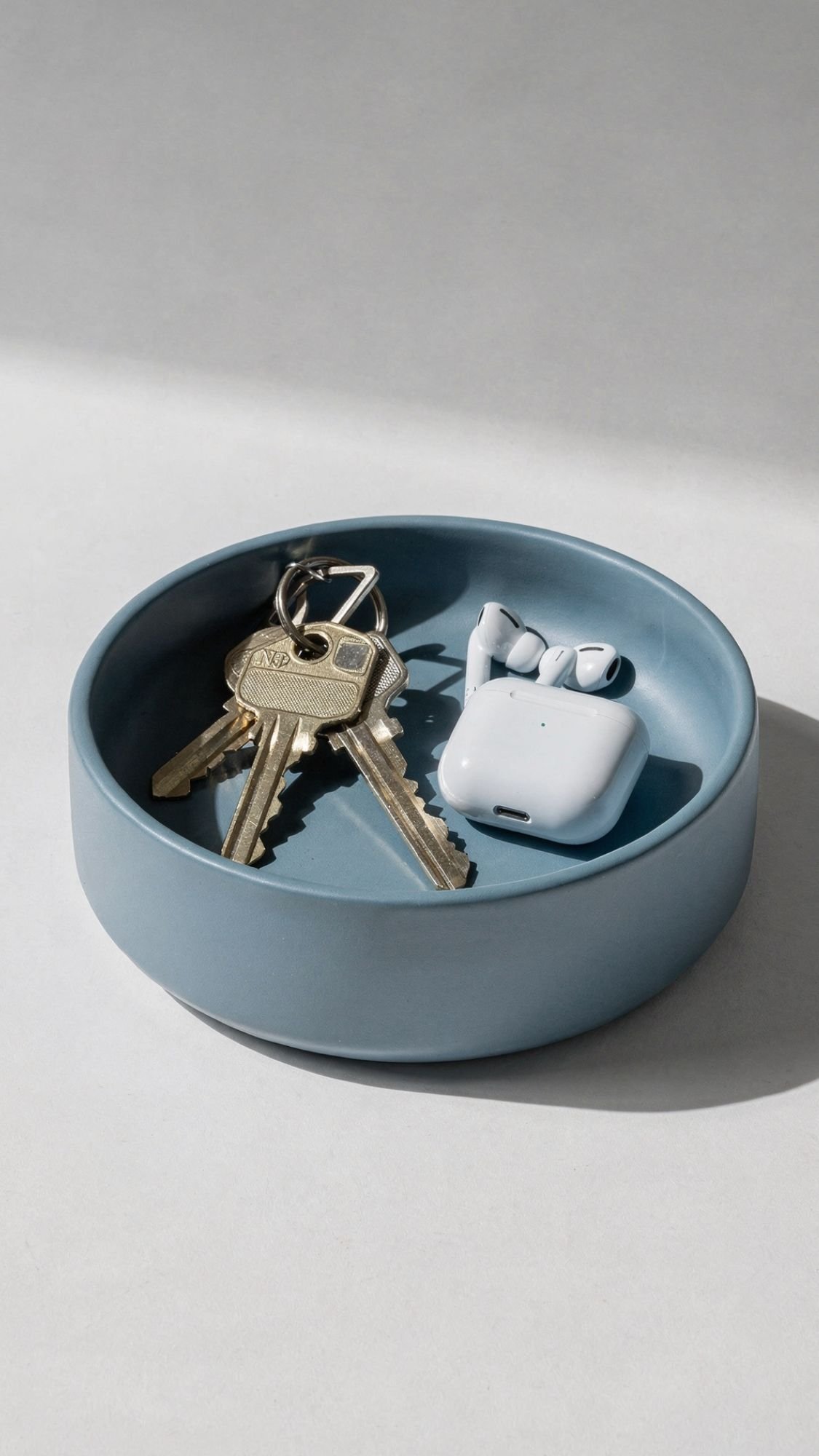 A light blue ceramic dish, perfect for useful gifts or best friend gifts, holds brass keys and wireless earbuds in a white charging case, casting soft shadows on a light surface.