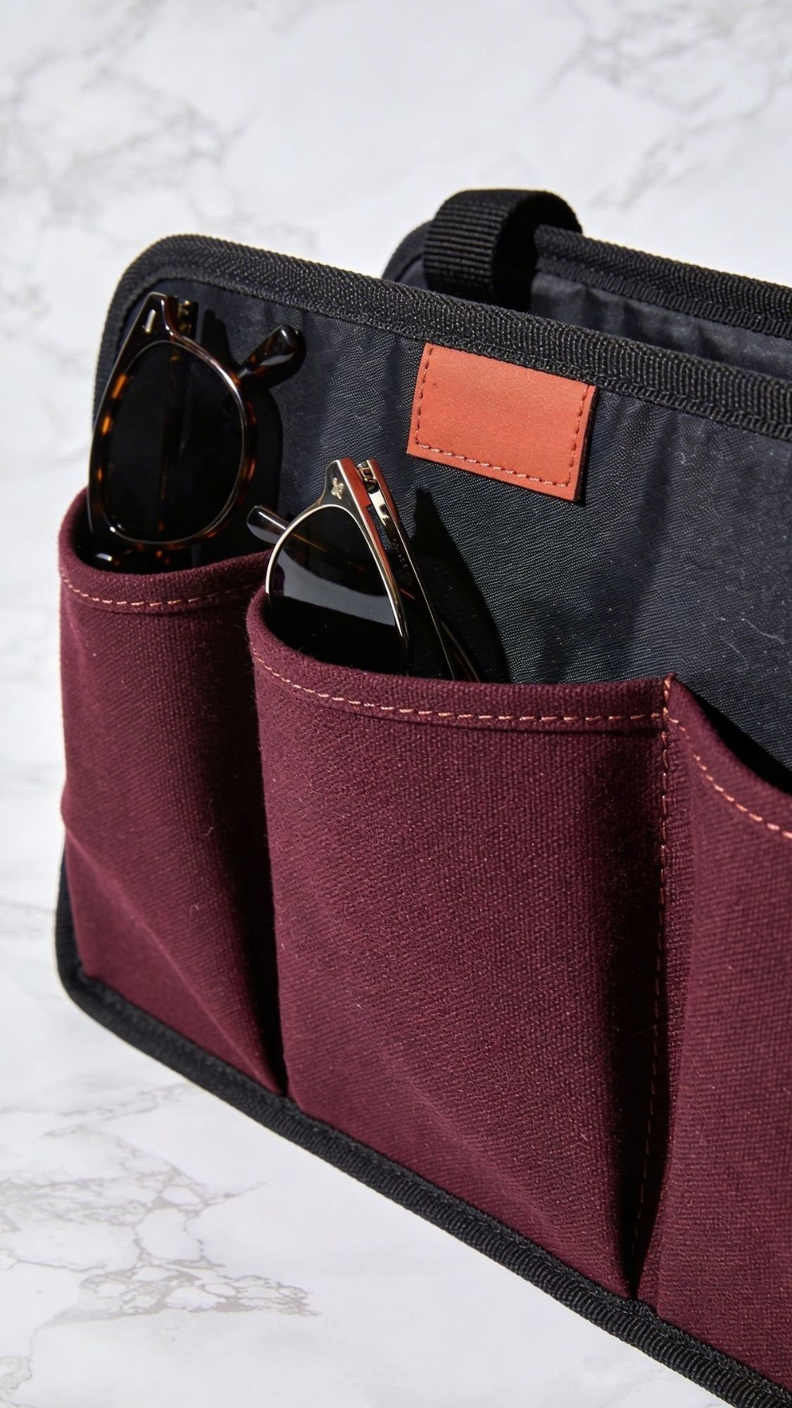 A close-up of a maroon fabric organizer with two front pockets holding a pair of sunglasses in each. The handmade gift features black trim and a small brown leather patch, perfect for best friend gifts, placed on a light marble surface.