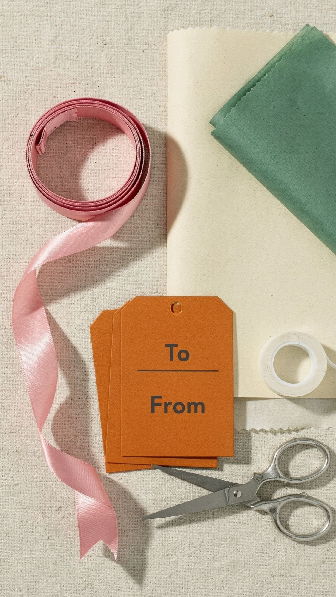 A flat lay of gift wrapping supplies—pink ribbon, green and cream tissue paper, brown To/From tags, clear tape, and silver scissors—perfect for wrapping unique gifts or handmade gift ideas for your best friend.