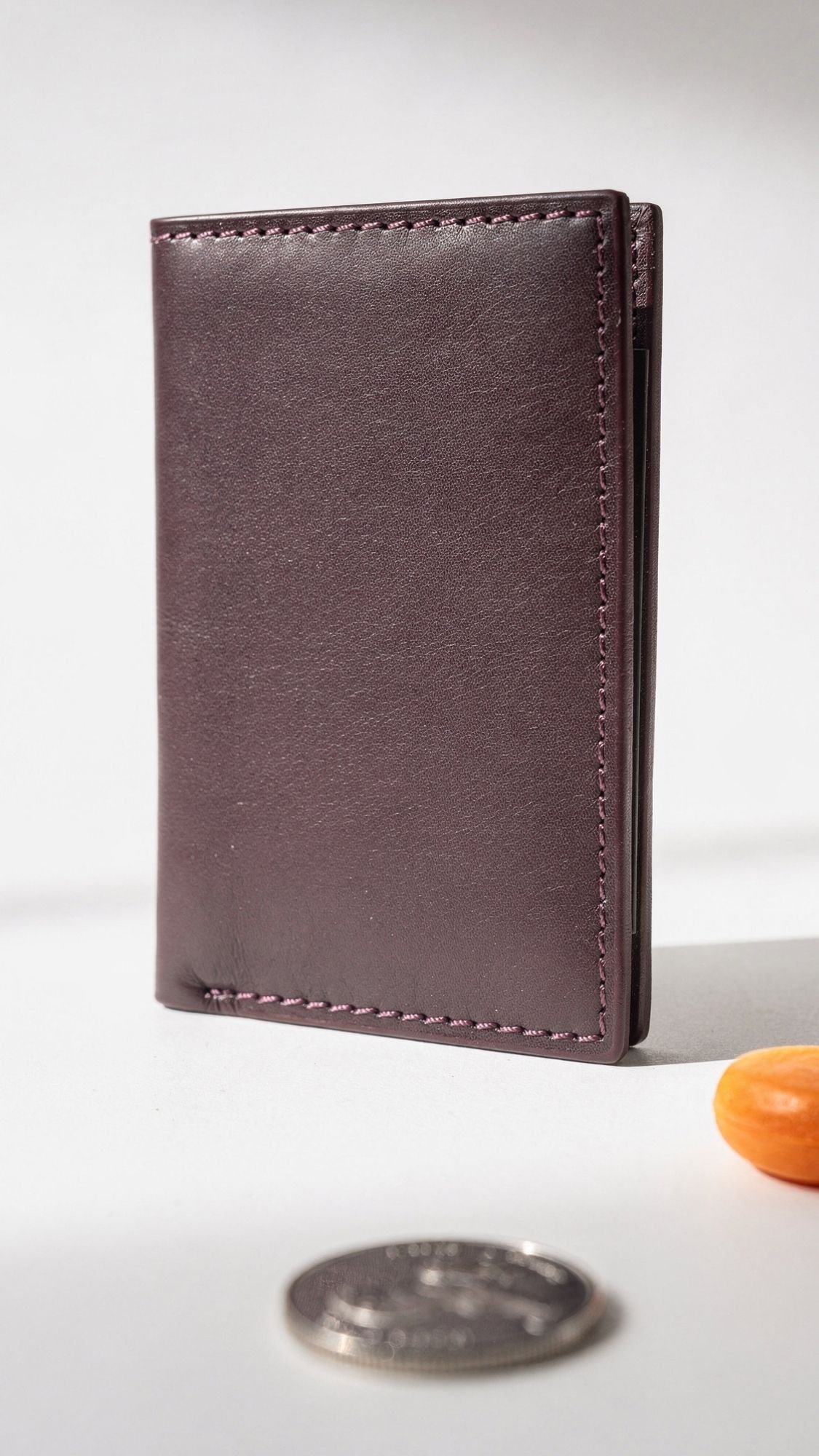 A dark brown leather wallet stands upright on a light surface next to a coin and an orange object, possibly a candy or button. With soft lighting and minimal shadows, it makes an ideal choice for handmade gifts for boyfriend.