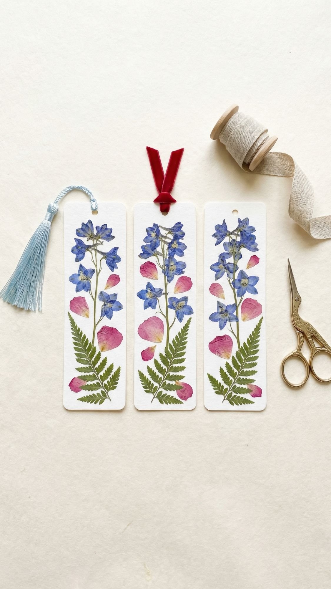 Three botanical bookmarks with pressed blue flowers, pink petals, and ferns are displayed. Each has a unique ribbon or tassel—perfect DIY presents or thoughtful gifts for Mother’s Day. Gold scissors and a beige spool of ribbon rest on a cream background.