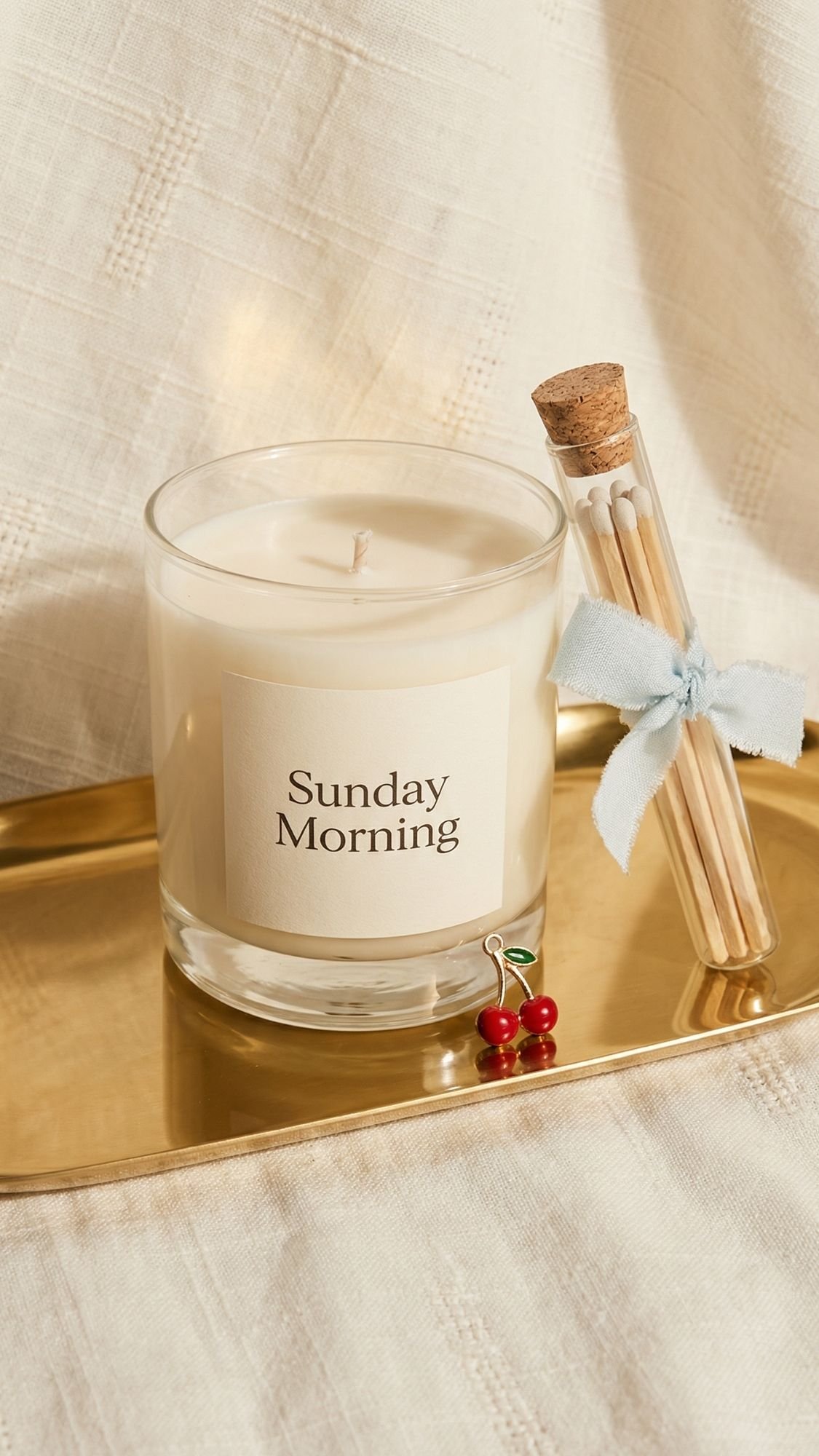 A glass candle labeled Sunday Morning sits on a gold tray next to a small glass vial of matches with a cork and blue bow, and a red cherry-shaped charm—perfect DIY presents or thoughtful Mother’s Day gifts—on textured cream-colored fabric.