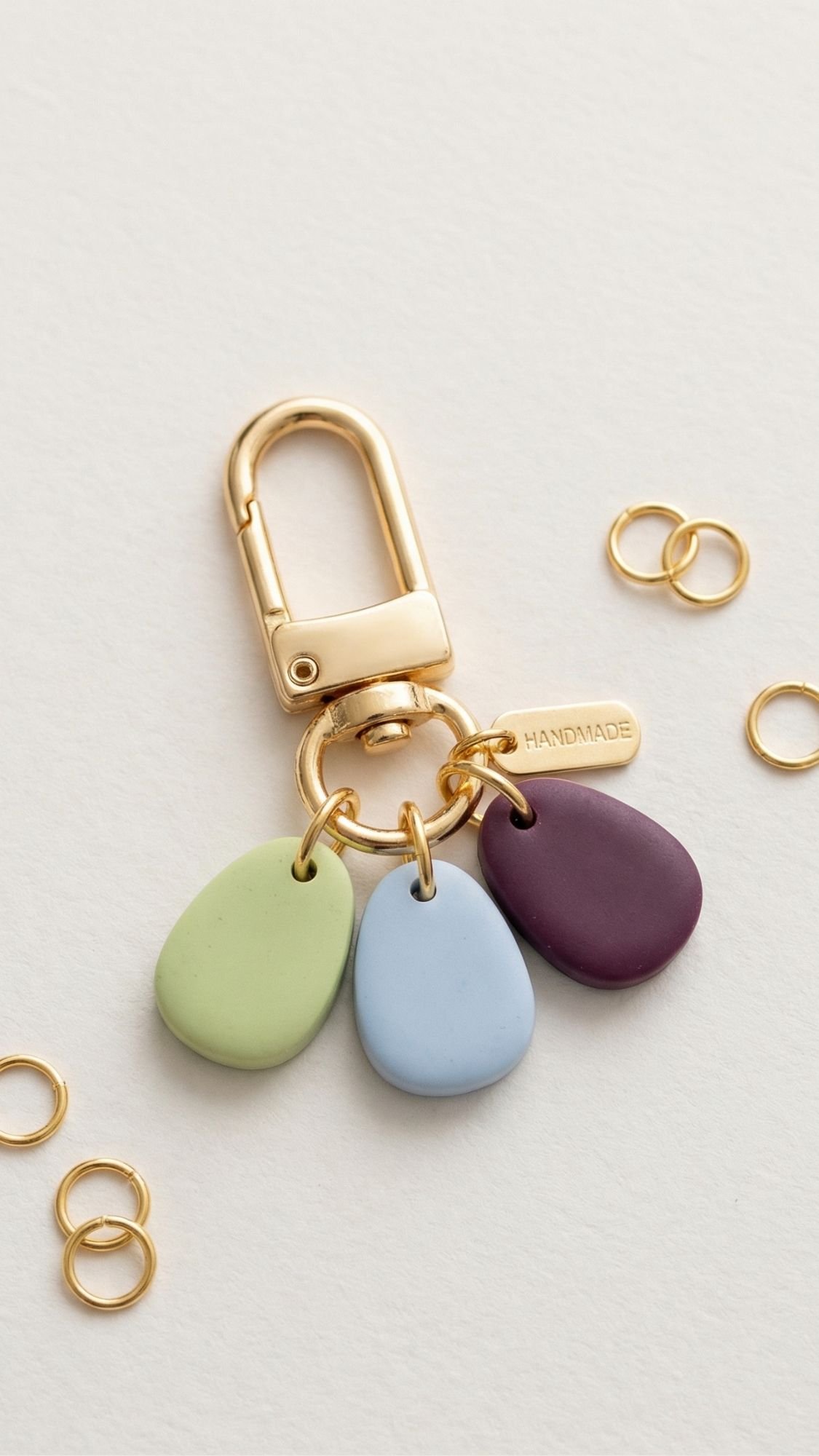 A gold keychain with a HANDMADE tag and three flat, pebble-shaped charms in green, blue, and purple—perfect as easy handmade gifts or thoughtful birthday gifts for best friend—lies on a light surface, surrounded by small gold rings.