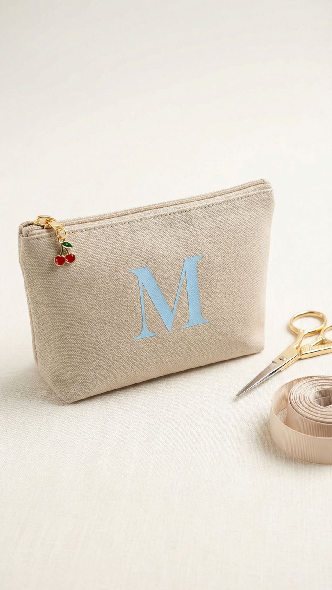 A beige fabric zipper pouch with a large blue letter M on the front—a charming choice for DIY Mother’s Day presents. The zipper features a cherry charm, while gold scissors and a beige measuring tape rest nearby on a light surface.