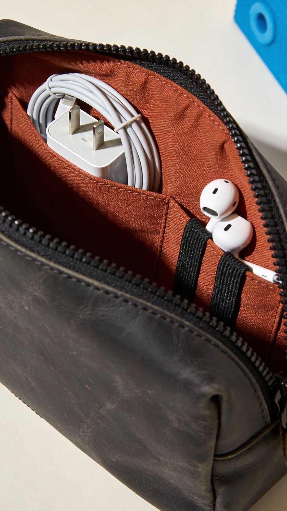A close-up of an open black leather pouch with a red interior, holding a coiled charging cable and wireless white earbuds—organized smartly in pockets—makes it one of the most useful gifts for boyfriend who loves staying organized.