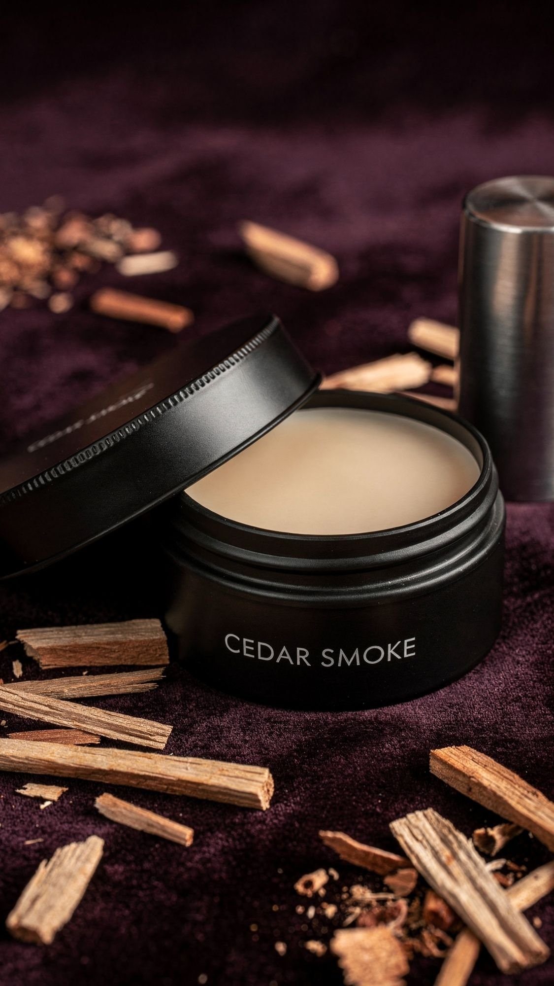 A black metal tin labeled Cedar Smoke containing a solid balm sits open on a dark purple surface, surrounded by scattered wood chips and a metallic cap nearby—an ideal choice for handmade boyfriend gifts.