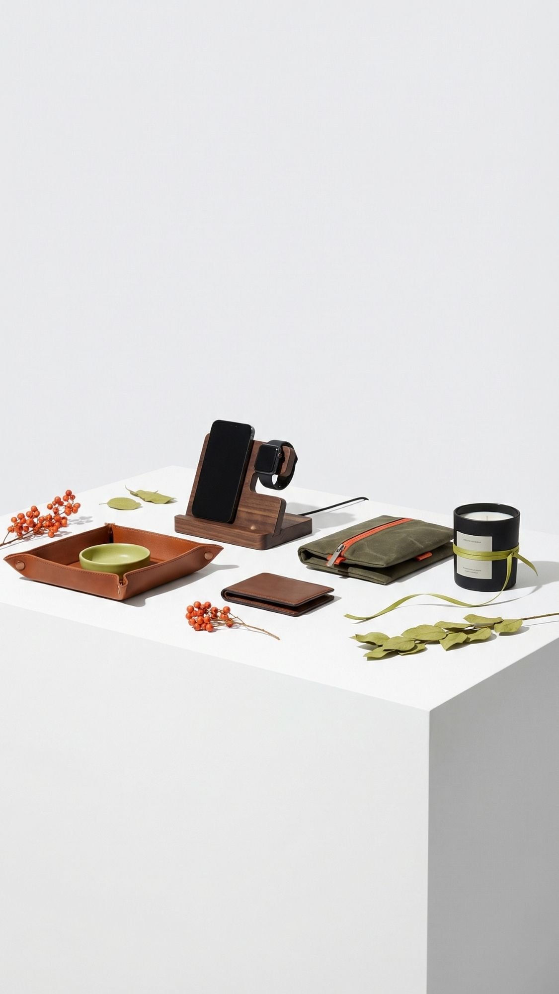 A modern white table displays a wooden phone and watch stand, a leather tray with a cup, coaster, zippered pouch, candle, and scattered dried leaves and berries—perfect handmade gifts for boyfriend or anyone who appreciates unique decor.