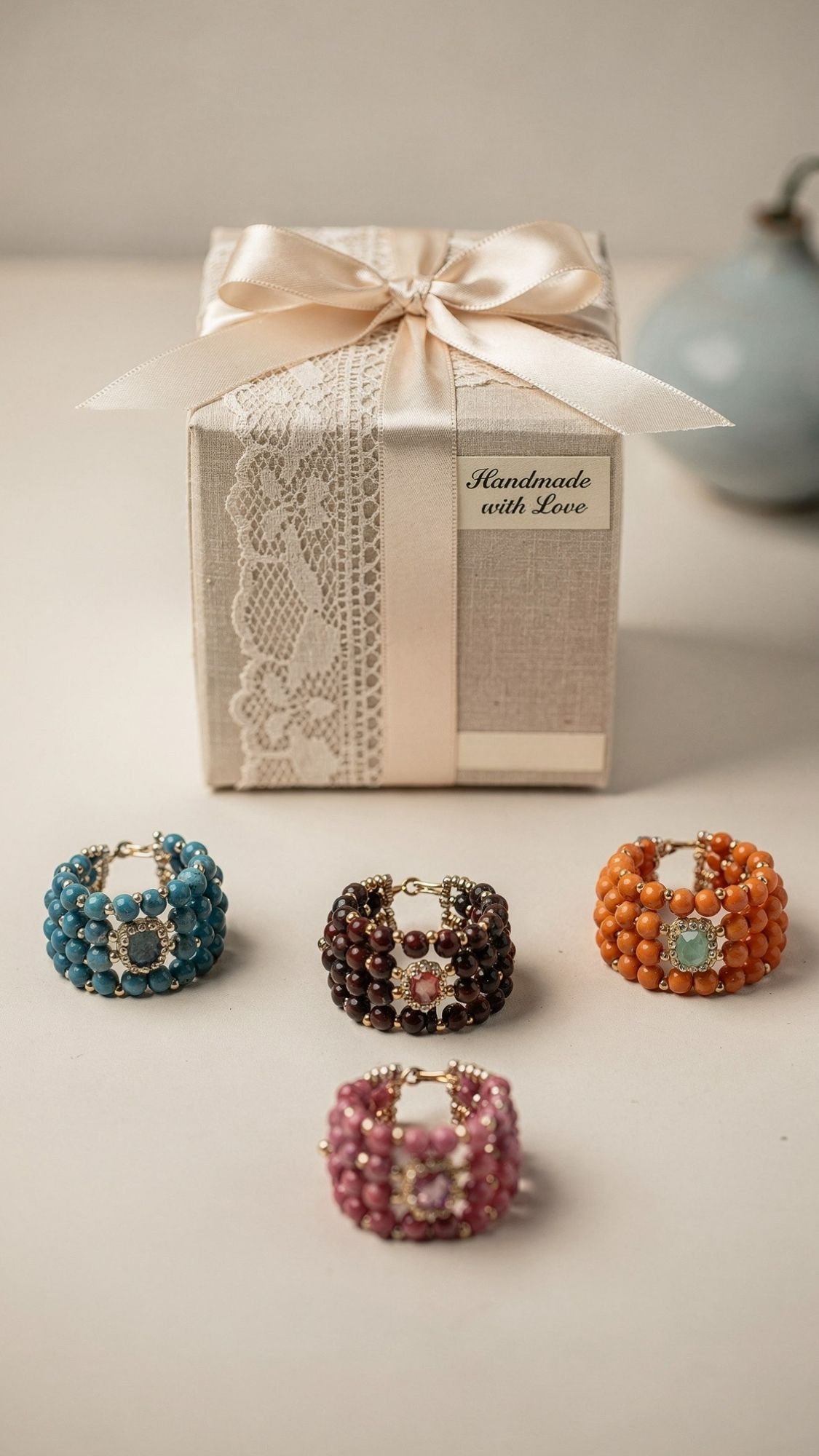 A gift box with a lace ribbon and a tag reading Handmade with Love sits behind four beaded rings in blue, brown, orange, and pink—each featuring a central gemstone—perfect as easy handmade gifts or thoughtful birthday gifts for best friend.