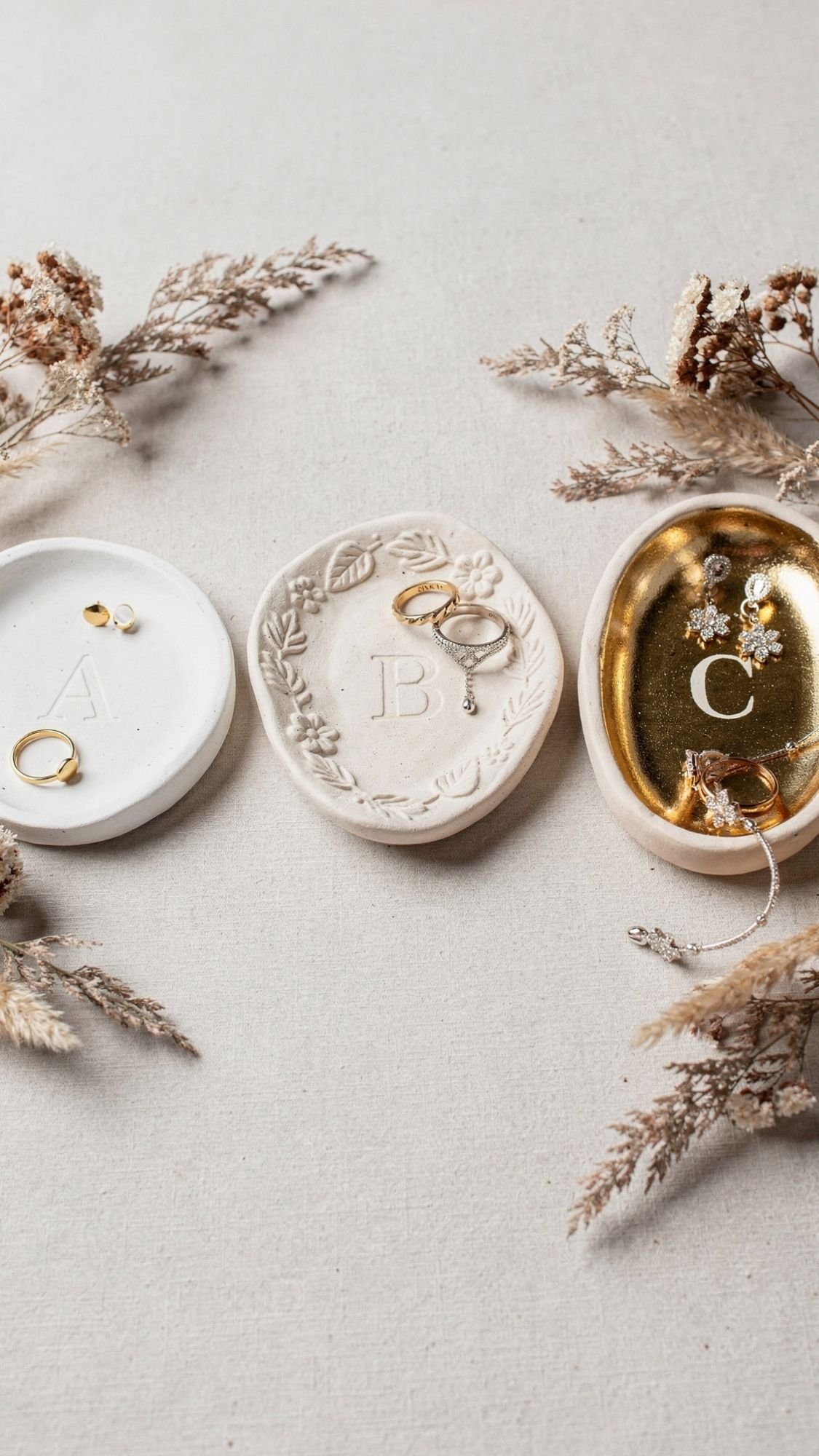 Three decorative ceramic dishes labeled A, B, and C display various gold and silver jewelry—perfect inspiration for homemade gifts. Dried beige foliage surrounds the dishes on a light fabric background.