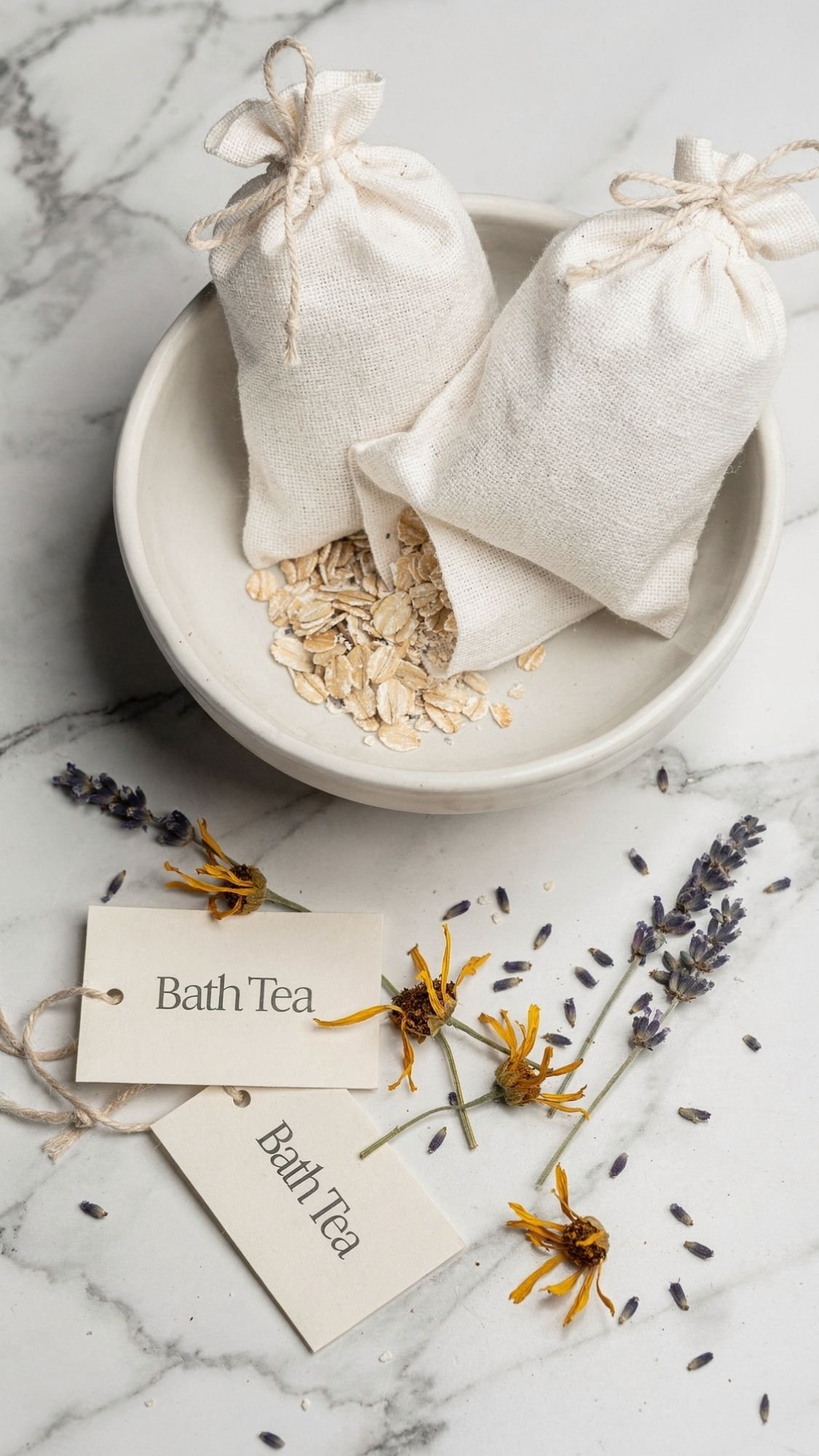 A marble surface with a white bowl holding oats and two cloth sachets, surrounded by dried lavender, yellow flowers, and two tags labeled Bath Tea—perfect for DIY spa gifts or an at home spa experience.