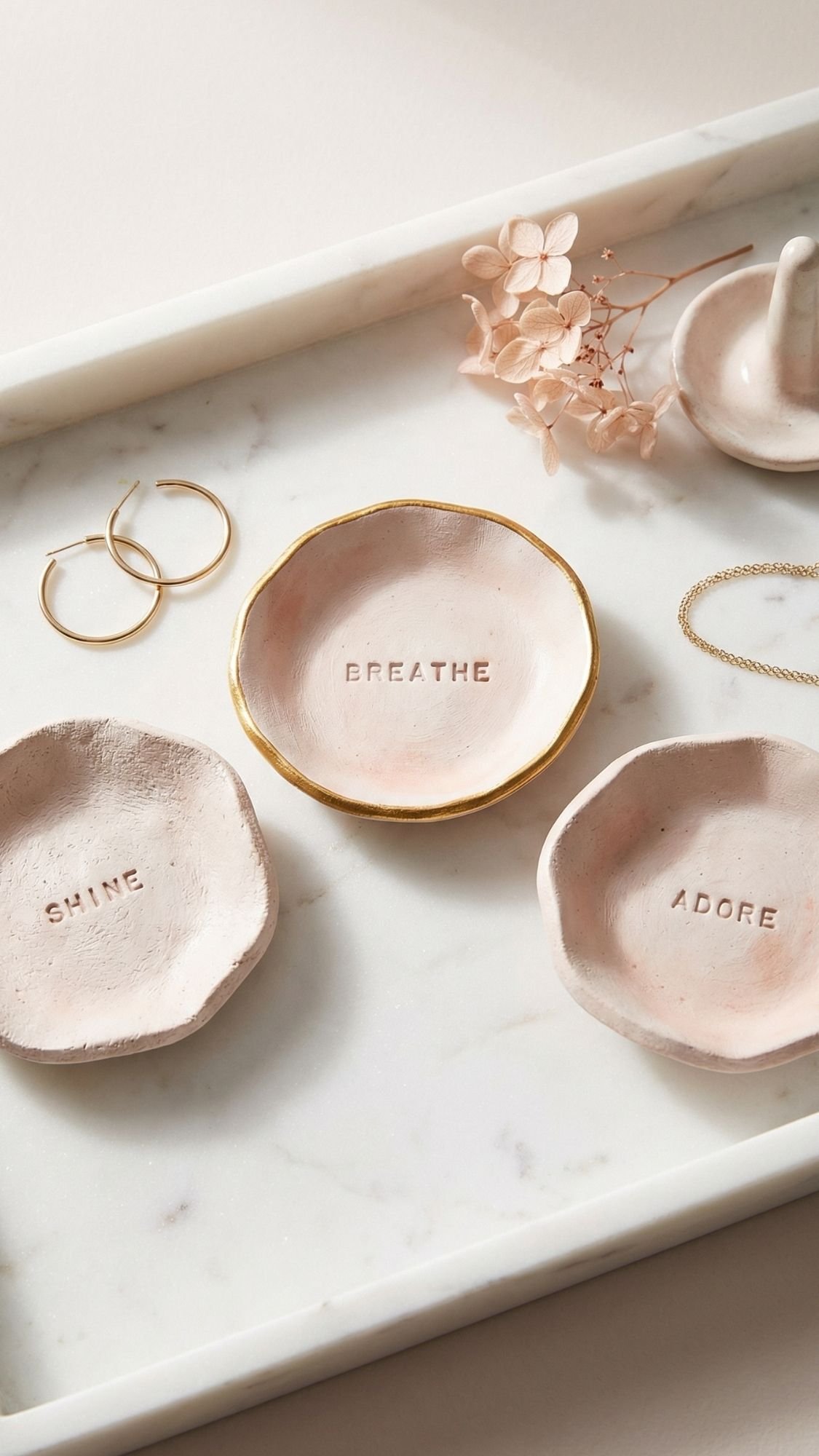 Three small, round ceramic trays on a marble surface, embossed with “SHINE,” “BREATHE,” and “ADORE.” Perfect as easy gifts or DIY presents for mom. Nearby sit hoop earrings, a delicate bracelet, dried flowers, and a ring holder.