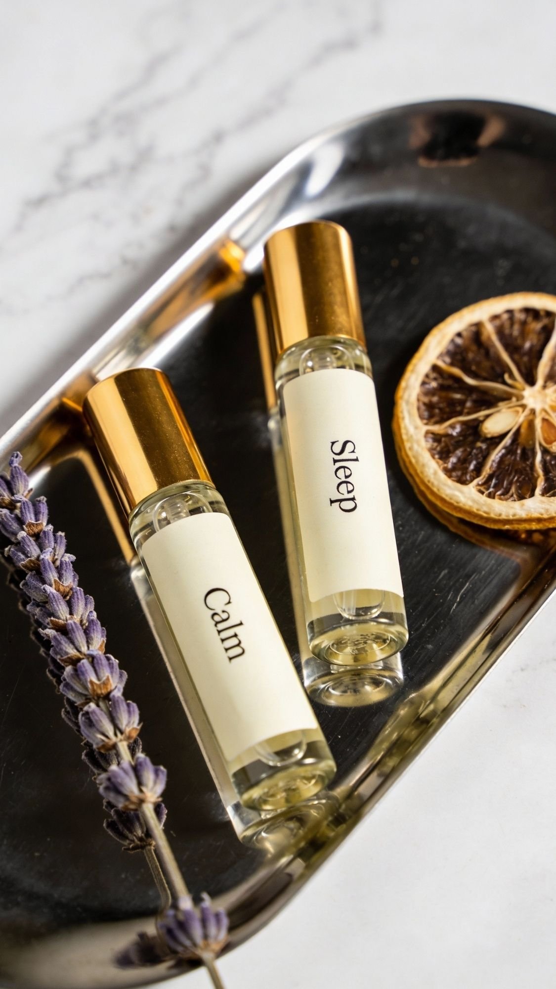Two glass roller bottles labeled Calm and Sleep with gold caps rest on a silver tray beside a dried orange slice and lavender sprig, making perfect DIY spa gifts or presents for mom on a relaxing spa day—all atop a white marble surface.