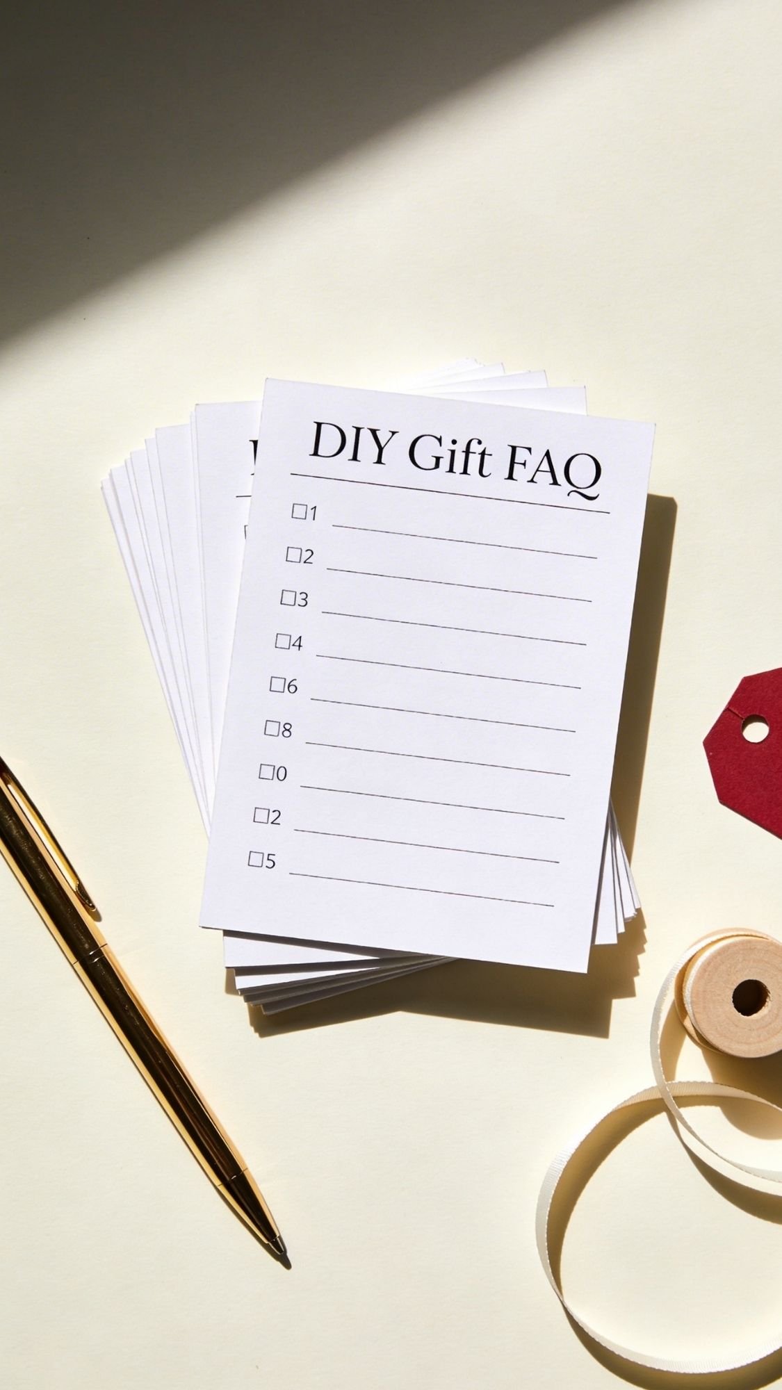 A stack of cards titled DIY Gift FAQ with a checklist and blank lines, placed beside a gold pen, spool of ribbon, and red gift tag on a light surface—perfect for jotting down Mother’s Day DIY Presents or creative Gift Ideas.