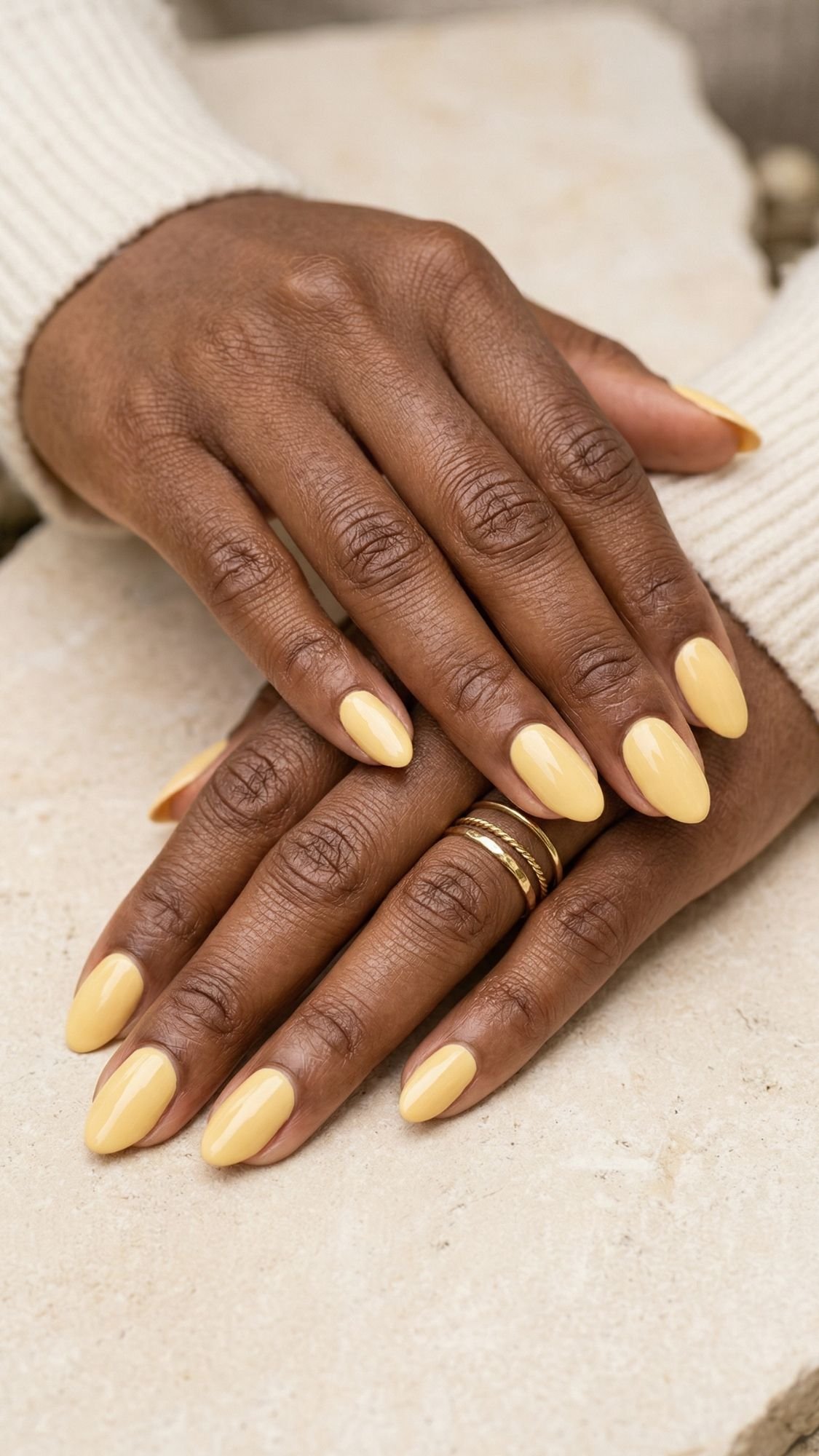A person with medium brown skin rests their hands on a light stone surface, displaying flattering almond-shaped nails painted in pale yellow polish. They wear a cream ribbed sweater and several thin gold rings.