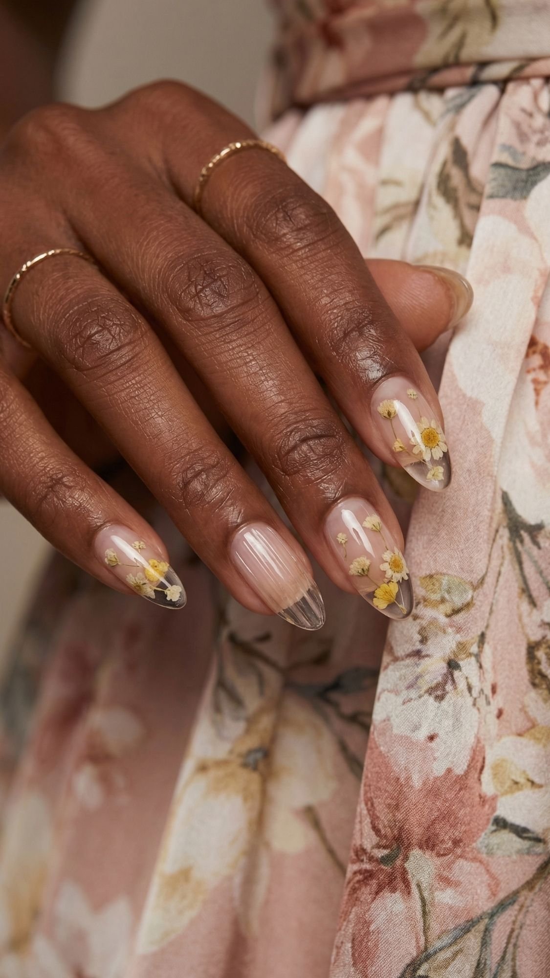 A hand with almond-shaped nails, each featuring a clear base and small yellow daisy designs—perfect for Summer Nails—rests on a soft pink floral dress. The person wears thin gold rings on three fingers.
