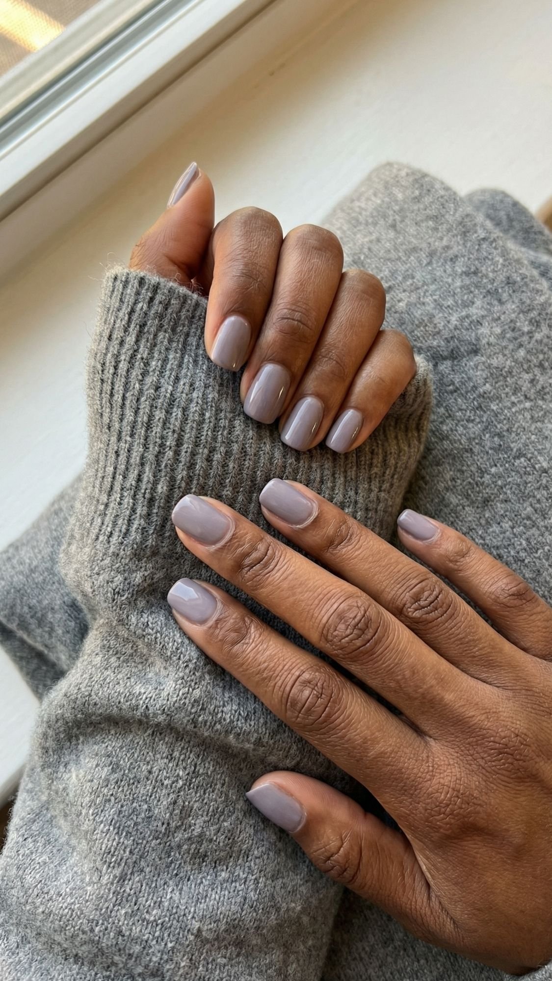 A person with medium brown skin wearing a cozy gray sweater shows off elegant nails, neatly manicured and painted in a soft, light lavender shade. Their short nails gently rest on each other near a window.
