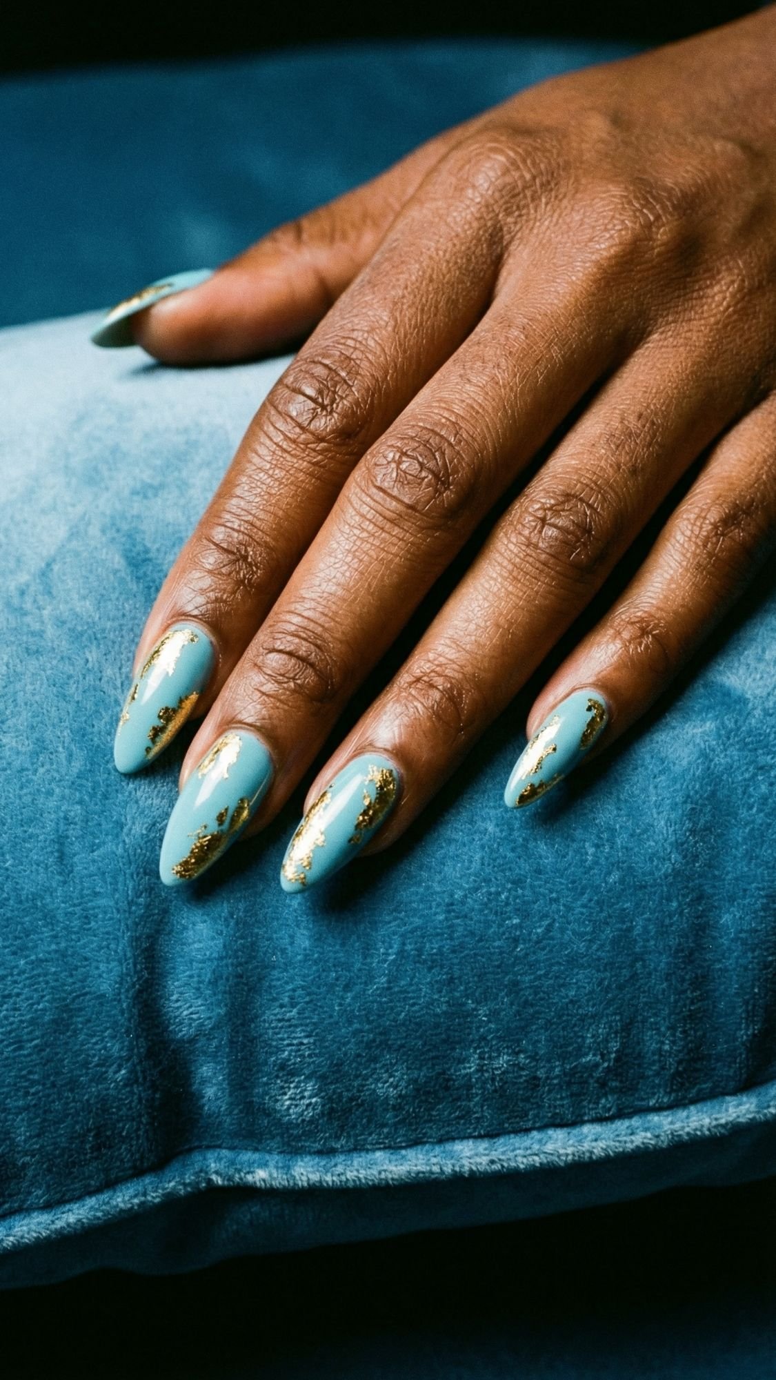 A hand with almond-shaped nails painted light blue with gold foil accents rests on a blue velvet cushion, showcasing Easter Holiday Nails perfect for those who love festive sparkle nails.
