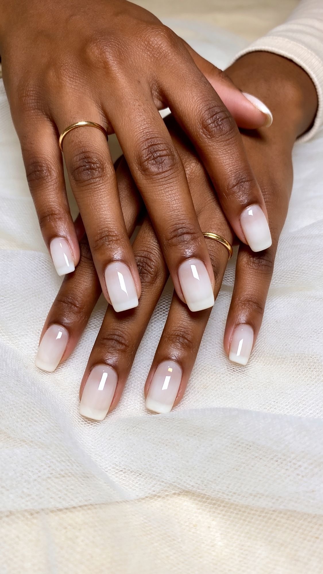 Two hands with medium-length square, glossy nude ombré nails—perfect wedding guest nails—resting on soft cream-colored fabric. The person wears simple gold rings on three fingers.