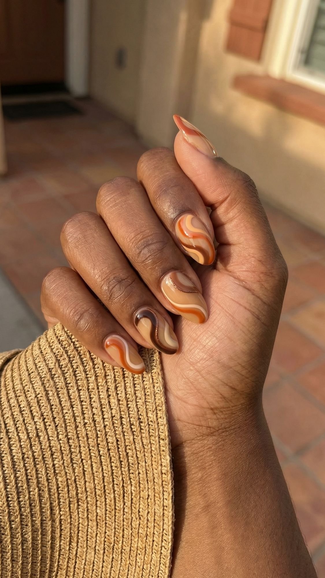 A hand with medium-length almond-shaped summer nails, featuring a brown, beige, and orange swirl pattern. The hand is held up outdoors, with a textured beige sleeve and a blurred background of a house and tiled floor.