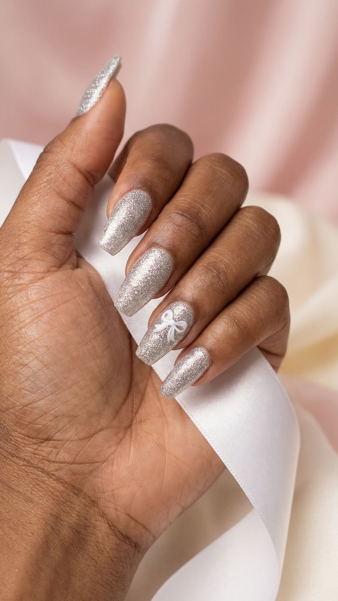 A hand with long, square-shaped nails painted silver with glitter. One nail features a white abstract design. Perfect for Easter Nails, the look is complemented by a white satin ribbon against a soft pink and cream background.