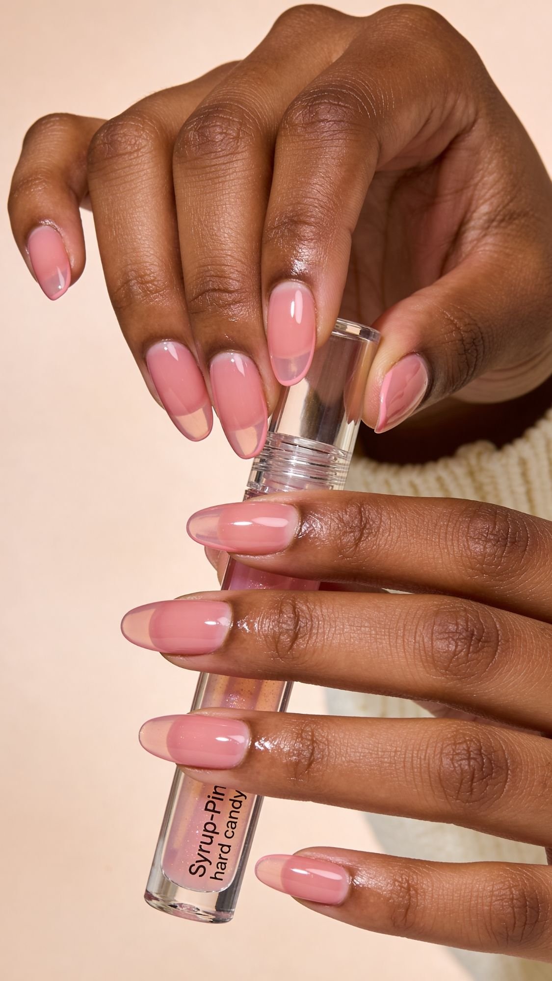 A close-up of two hands with glossy, pink, almond nails holding a clear tube labeled Syrup-Pit hand candy against a neutral background—showcasing one of the most flattering nail designs.