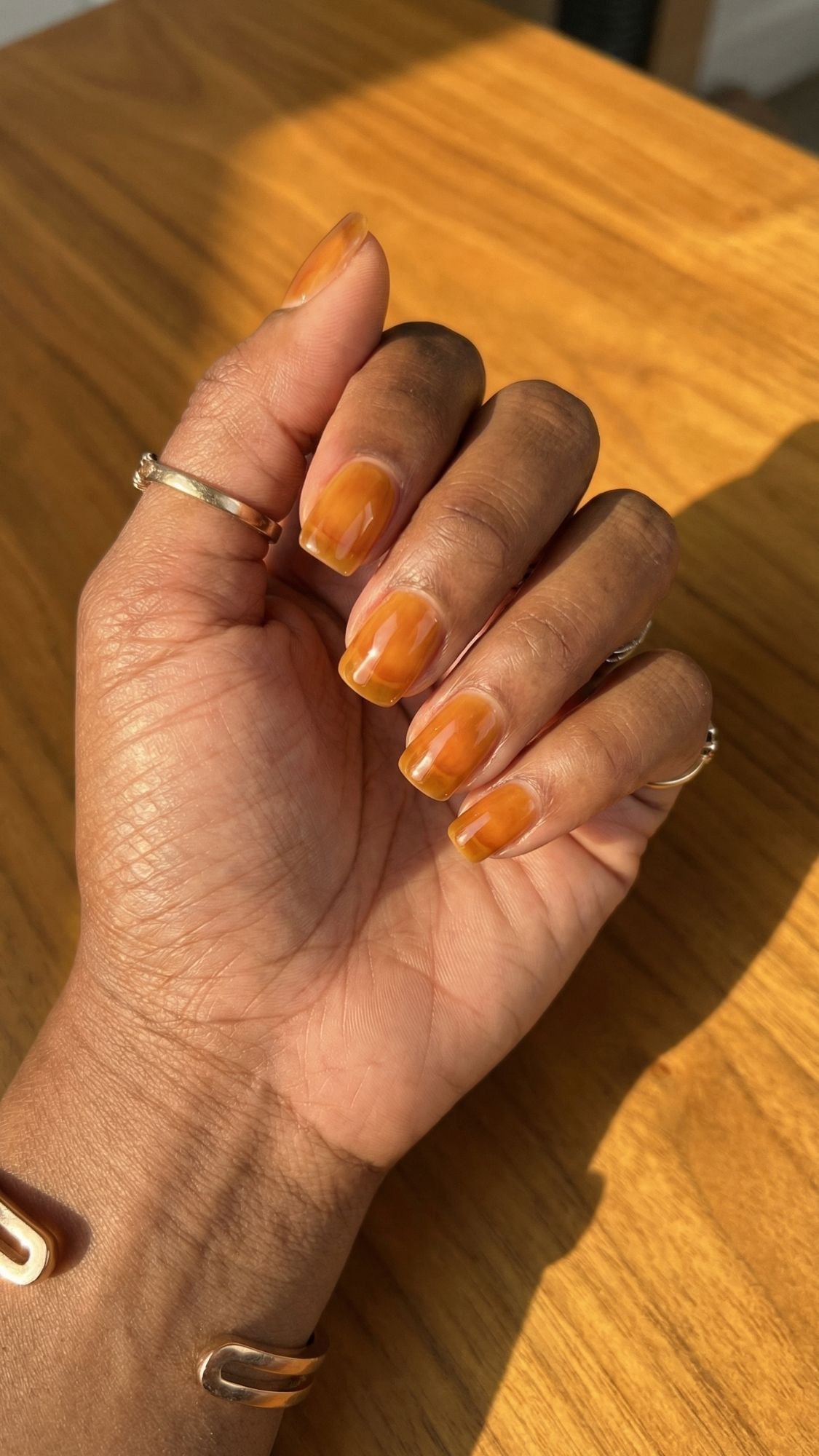 A hand with brown skin and gold rings displays neatly manicured nails painted in a glossy, amber-orange shade with subtle marble-like patterns, capturing the essence of Clean Girl Nails for a polished everyday look on a wooden surface in sunlight.